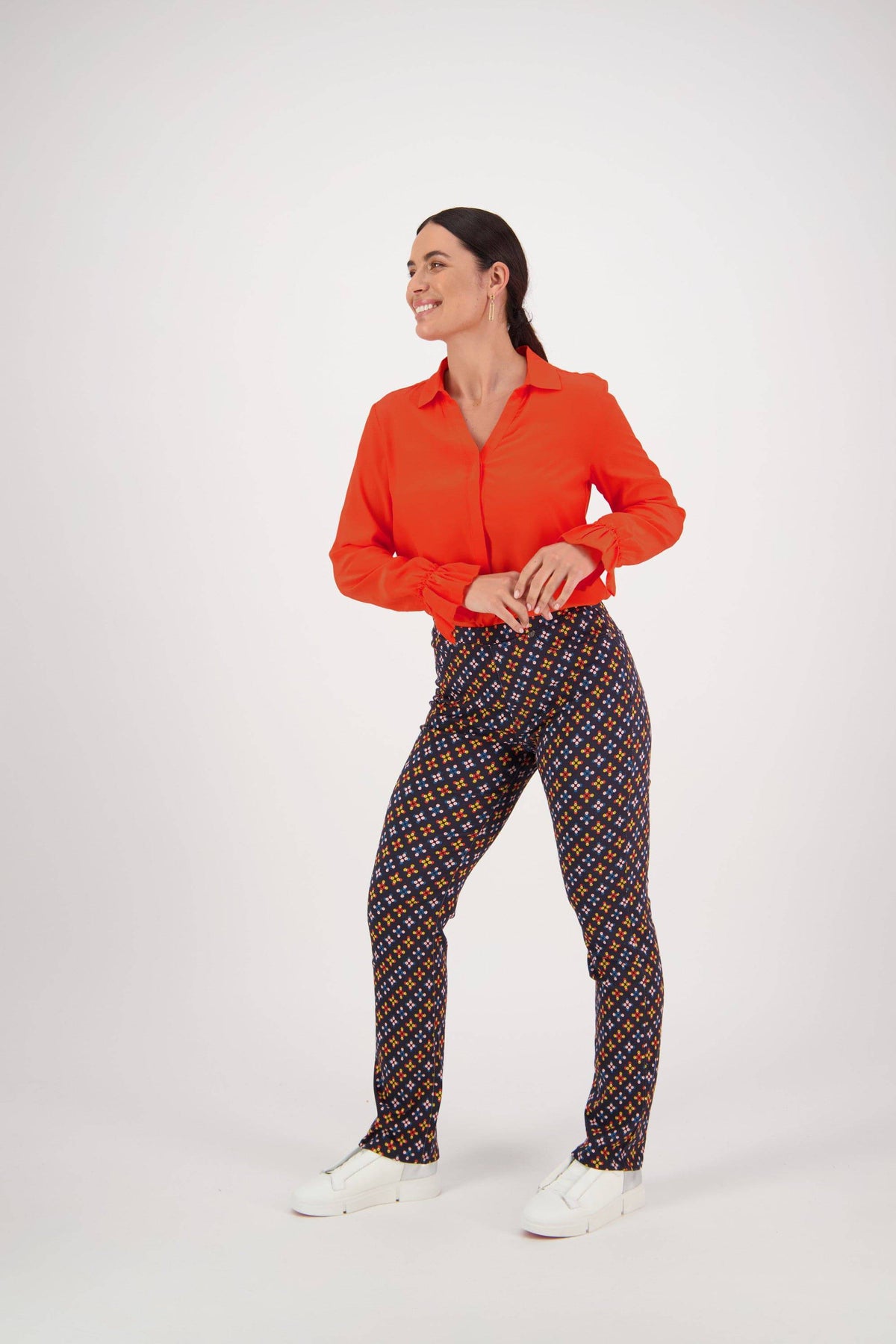 A woman stands smiling in a bright orange long-sleeve shirt, Vassalli Bay Pull On - Stellar slim leg pants with a colorful print, and white sneakers, set against a plain white background.