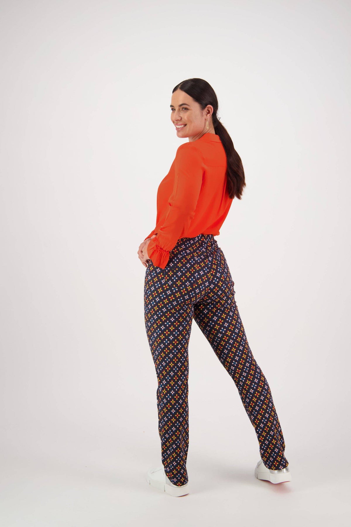 A woman with a ponytail smiles over her shoulder, wearing a bright orange blouse, Vassalli Bay Pull On - Stellar pants in patterned navy blue, and white shoes. The background is plain white.
