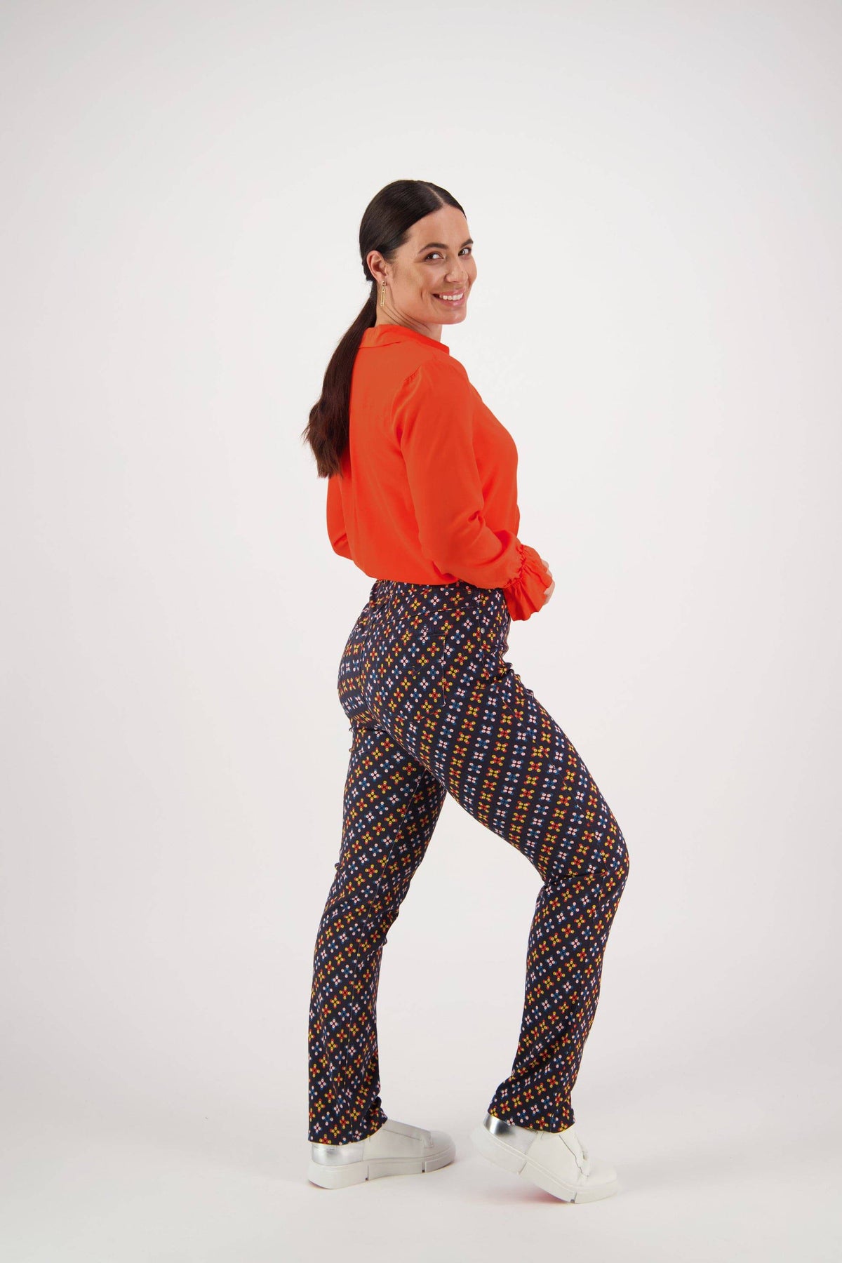 A woman smiles while facing sideways, looking at the camera. She wears a bright orange long-sleeve top, Vassalli Bay Pull On - Stellar pants, and white sneakers against a plain white background.