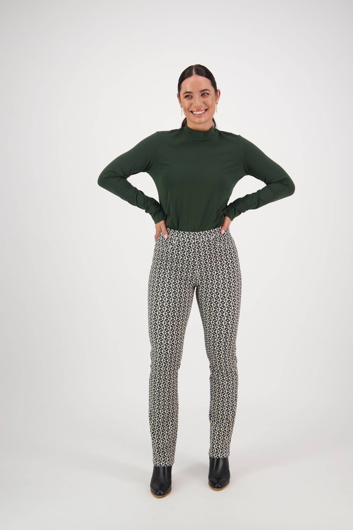 A woman stands smiling with hands on hips, wearing a dark green long-sleeve top, Vassalli's Bay Pull On - Vault patterned high-waisted pants, and black shoes, against a plain white background.