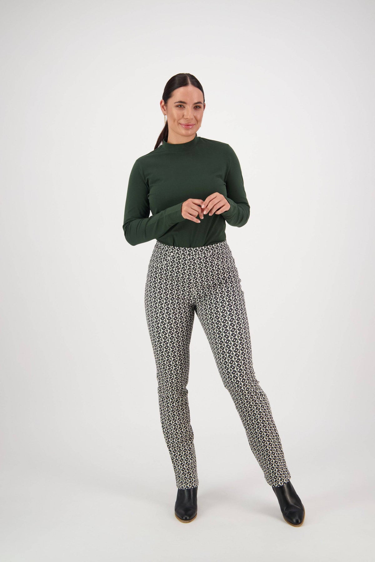 A woman stands against a plain white background wearing a dark green long-sleeve top and Vassalli Bay Pull On - Vault pants—black-and-white patterned slim leg pull-ons in stretch fabric. She smiles, hands loosely clasped, and wears black ankle boots.