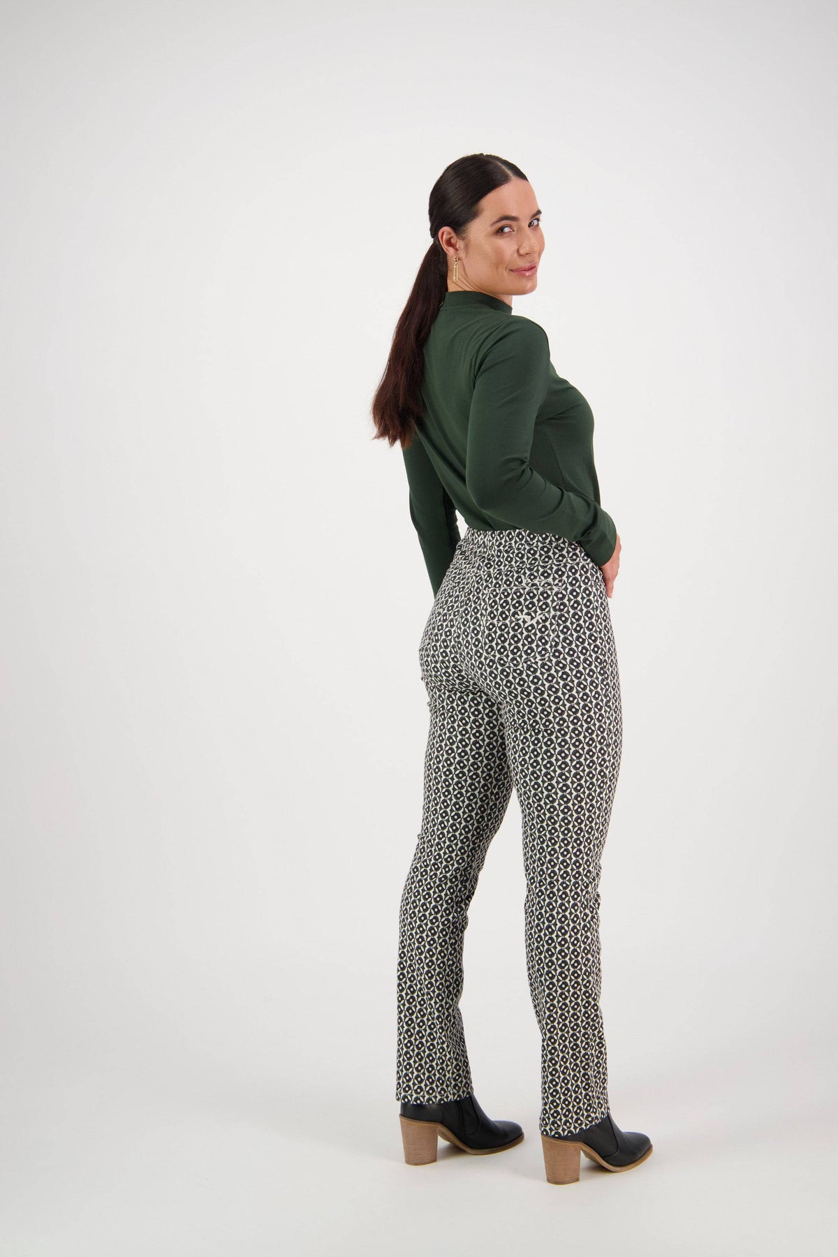 A woman with long dark hair in a ponytail wears a dark green long-sleeve top, Vassalli Bay Pull On - Vault pants, and black ankle boots. She stands turned slightly to the side, smiling at the camera against a plain white background.