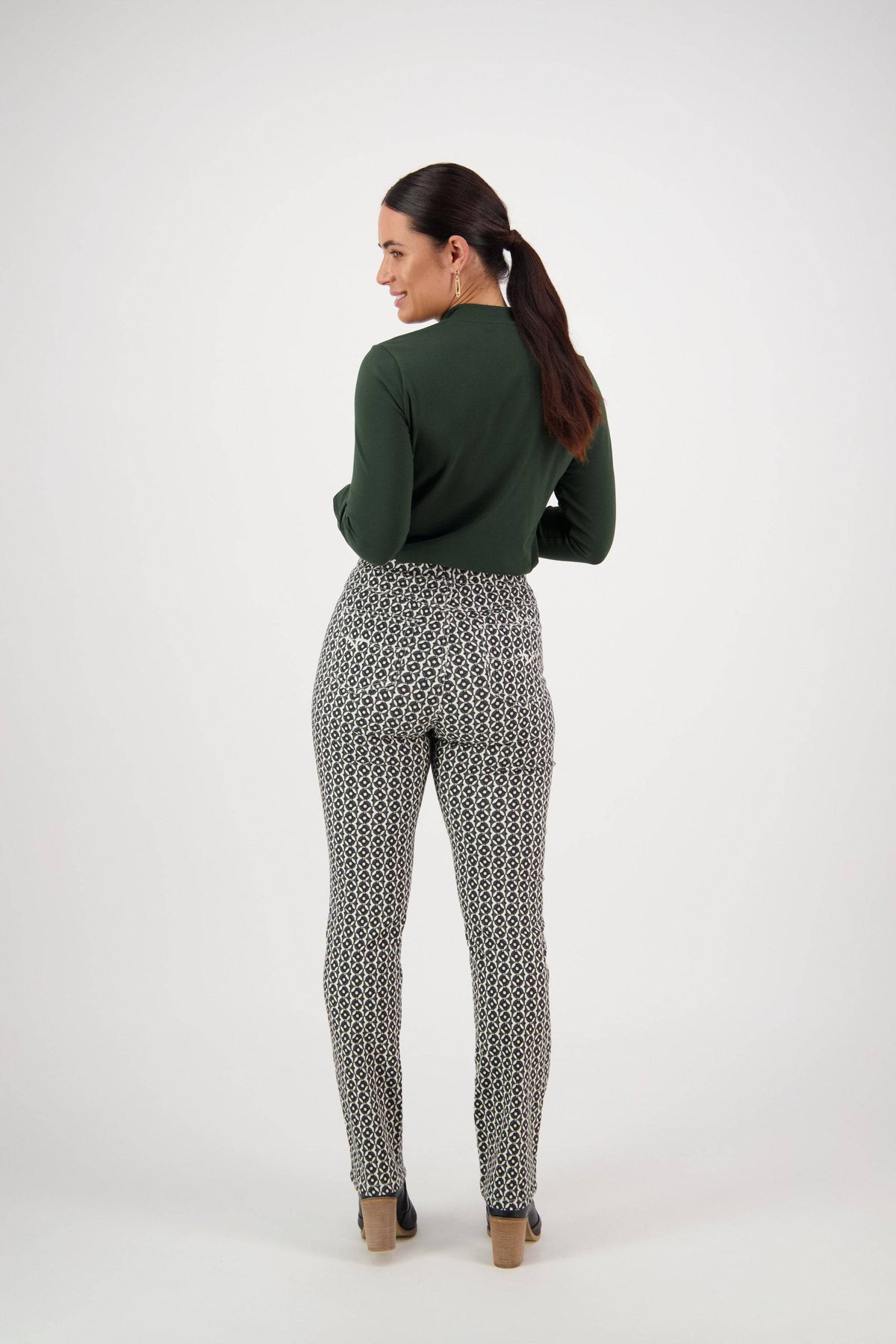 A woman with long dark hair in a ponytail faces away, wearing a dark green long-sleeve top and Vassalli Bay Pull On - Vault slim leg pants, paired with beige heels against a plain white background.
