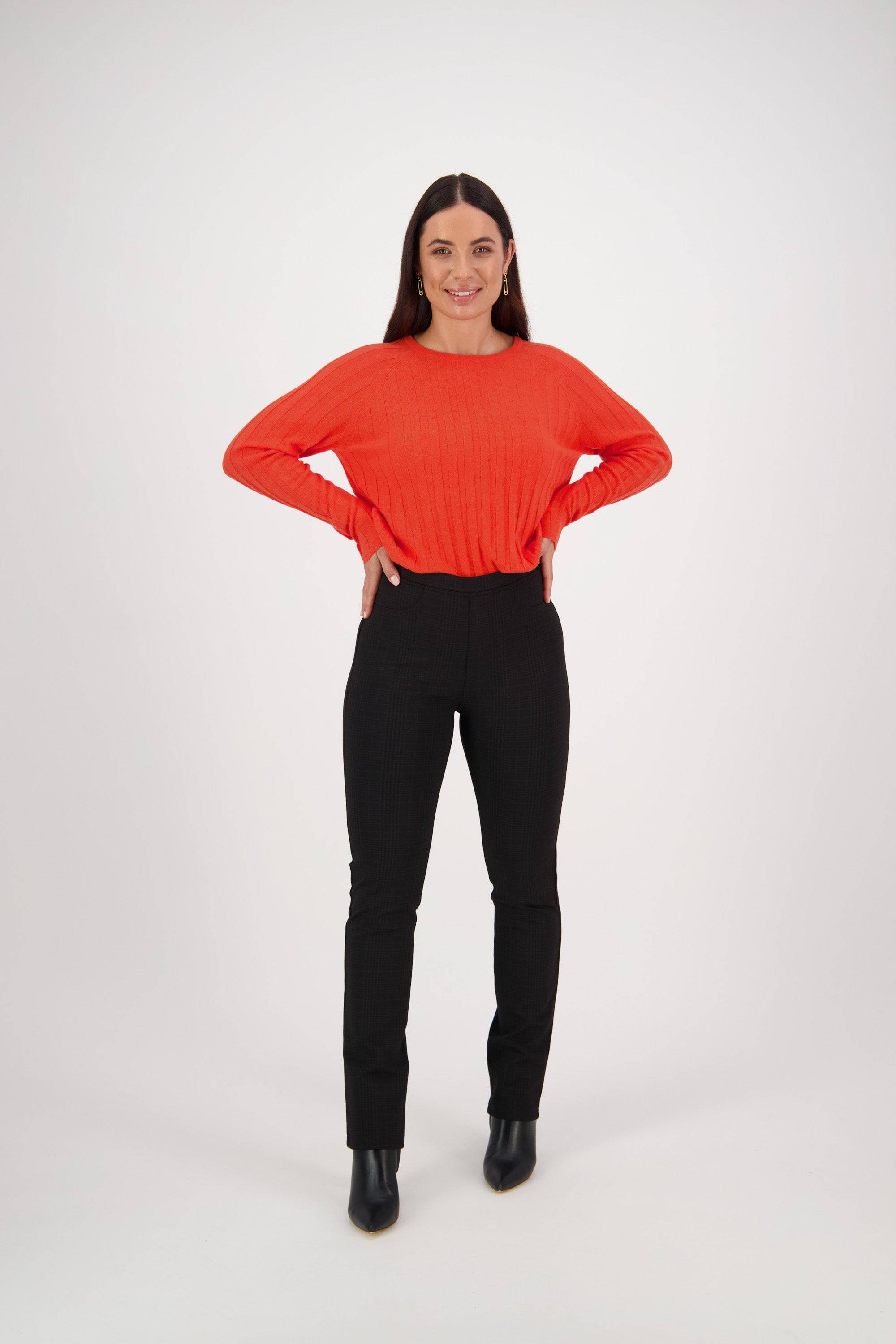 A woman smiles with hands on hips, wearing the Bay Ponti Pull On - Albert pants by Vassalli in black, a bright orange sweater, and black ankle boots, standing against a plain white background.