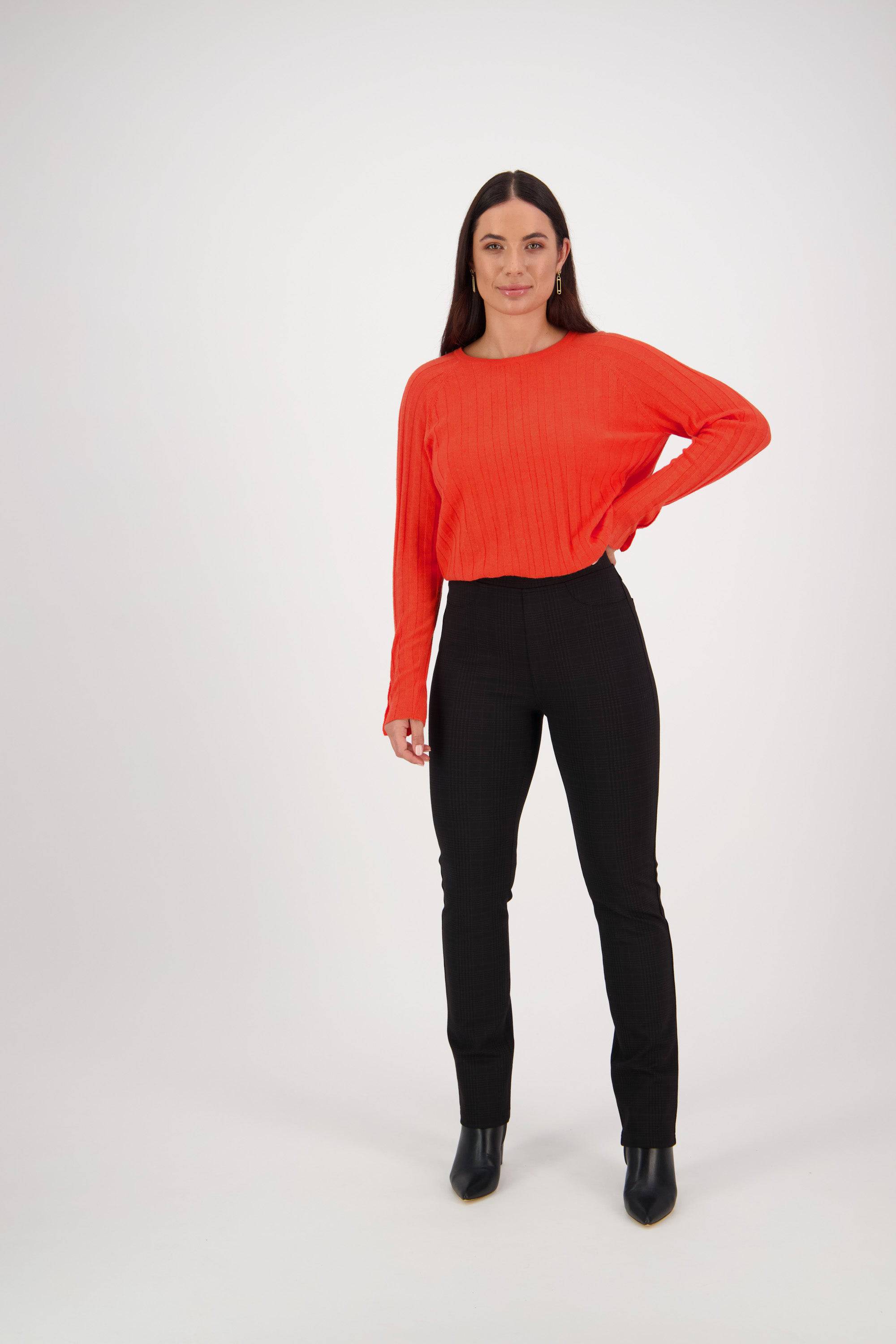 A woman with long dark hair stands against a white background, wearing the Vassalli Bay Ponti Pull On - Albert and black ankle boots. She looks at the camera with a slight smile in a bright orange long-sleeve top.
