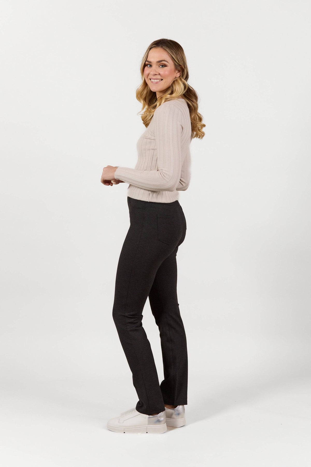A young woman with long, wavy blonde hair stands sideways, smiling. She wears Vassalli’s Bay Ponti Pull On pants in charcoal with a beige sweater and white sneakers against a plain white background.