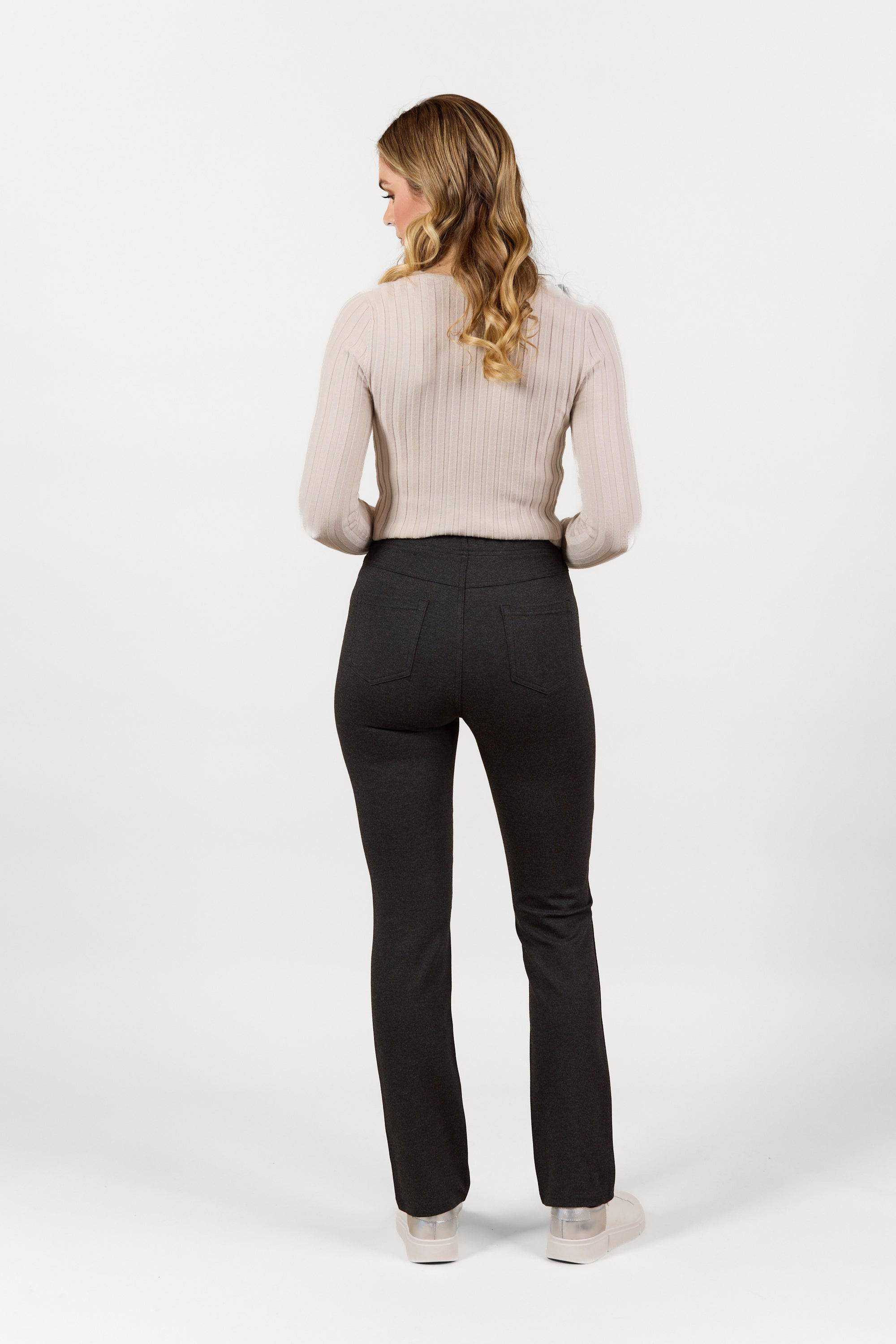 A woman with long wavy hair, wearing the Vassalli Bay Ponti Pull On charcoal pants and a light beige ribbed sweater, stands facing away from the camera against a plain white background.