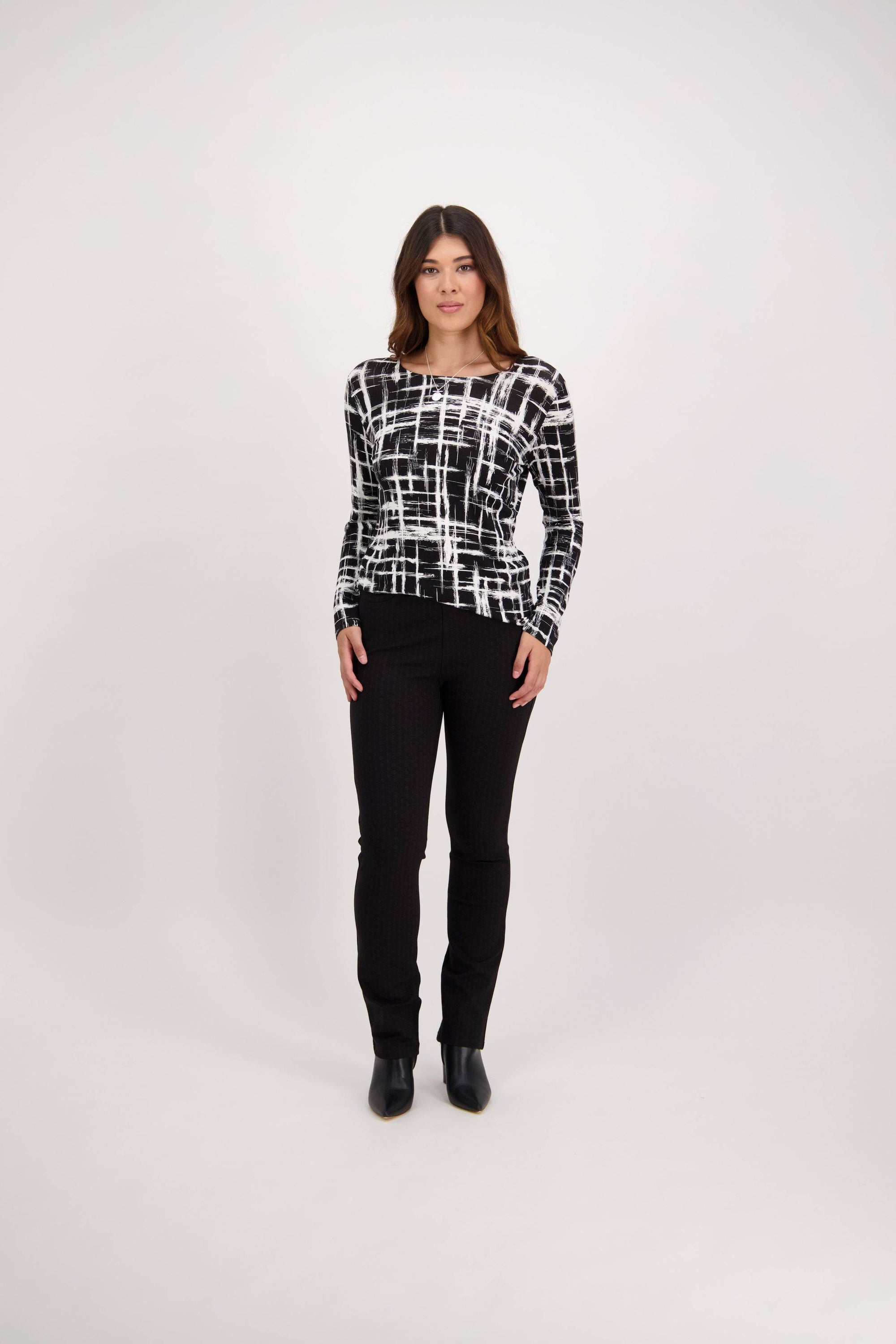 A woman with shoulder-length hair and a neutral expression wears Vassalli's Bay Ponti Pull On - Colombia high-rise slim leg pants, a grid-patterned top, and black ankle boots against a plain white background.