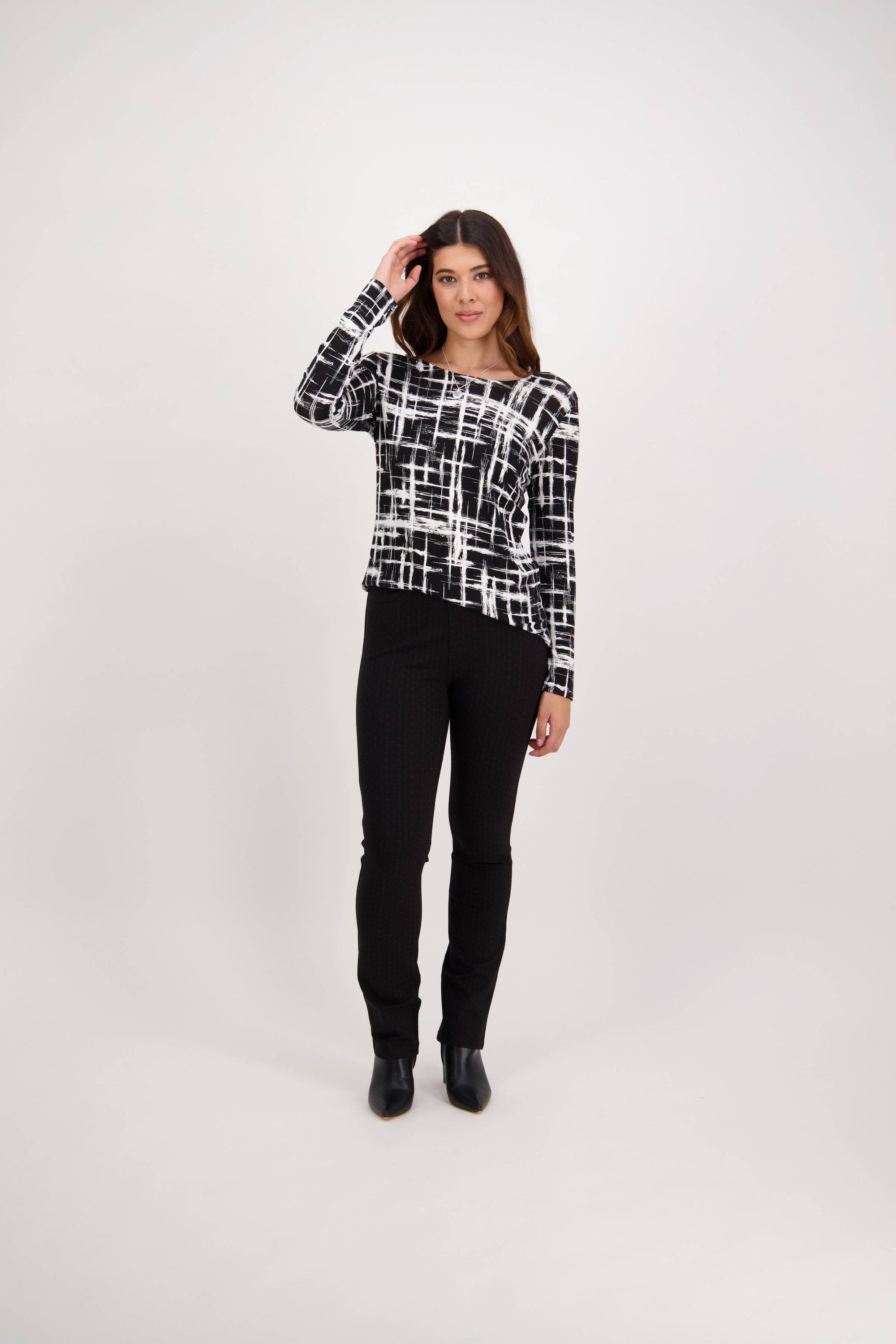 A woman stands against a plain white background, wearing a black and white grid-patterned long-sleeve top, Vassalli Bay Ponti Pull On - Colombia pants, and black ankle boots. She has one hand in her hair and a neutral expression.