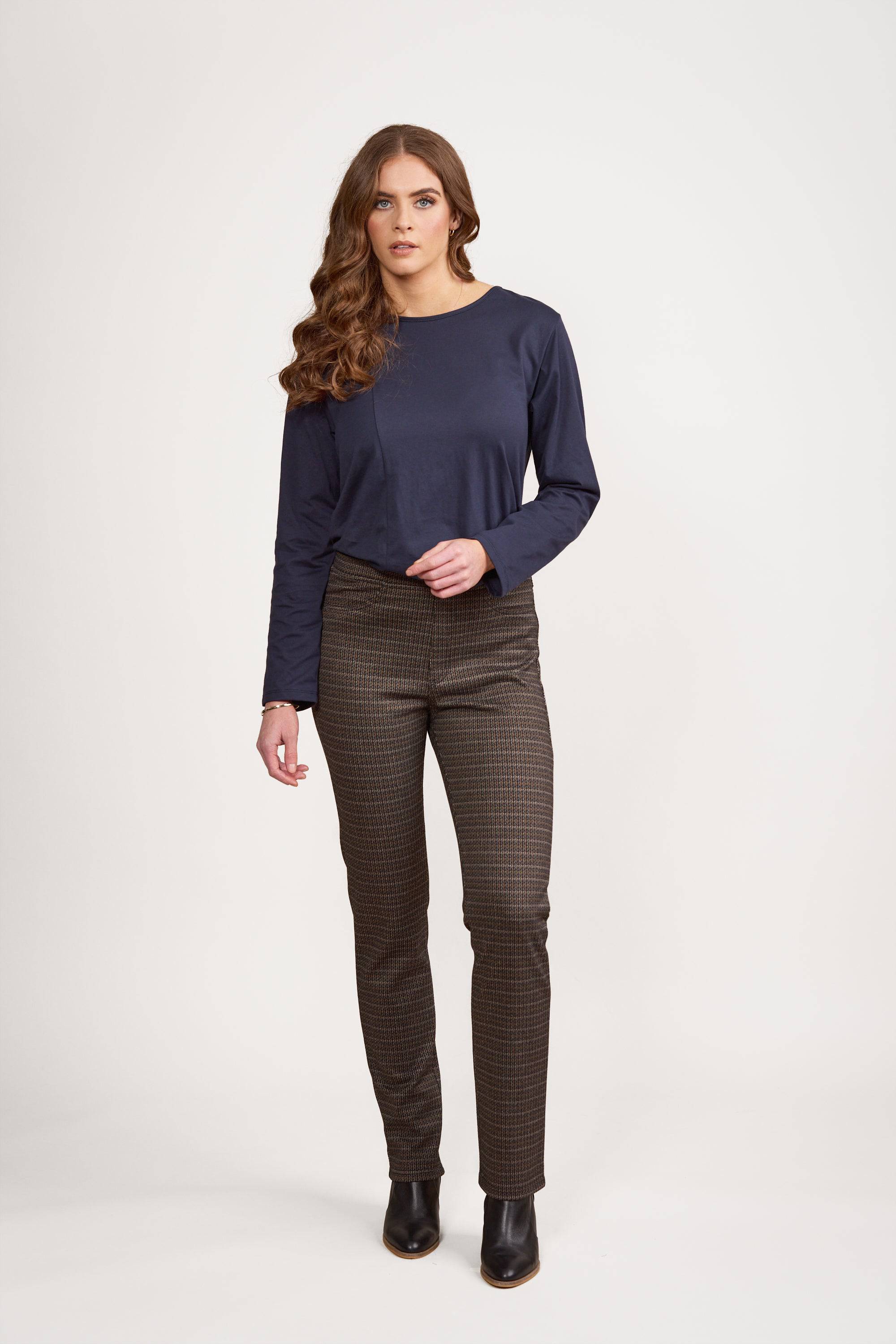 A woman with long wavy brown hair wears a navy long-sleeve top, Vassalli Bay Knit Pull On - Sutton pants, and black ankle boots, standing against a plain white background.