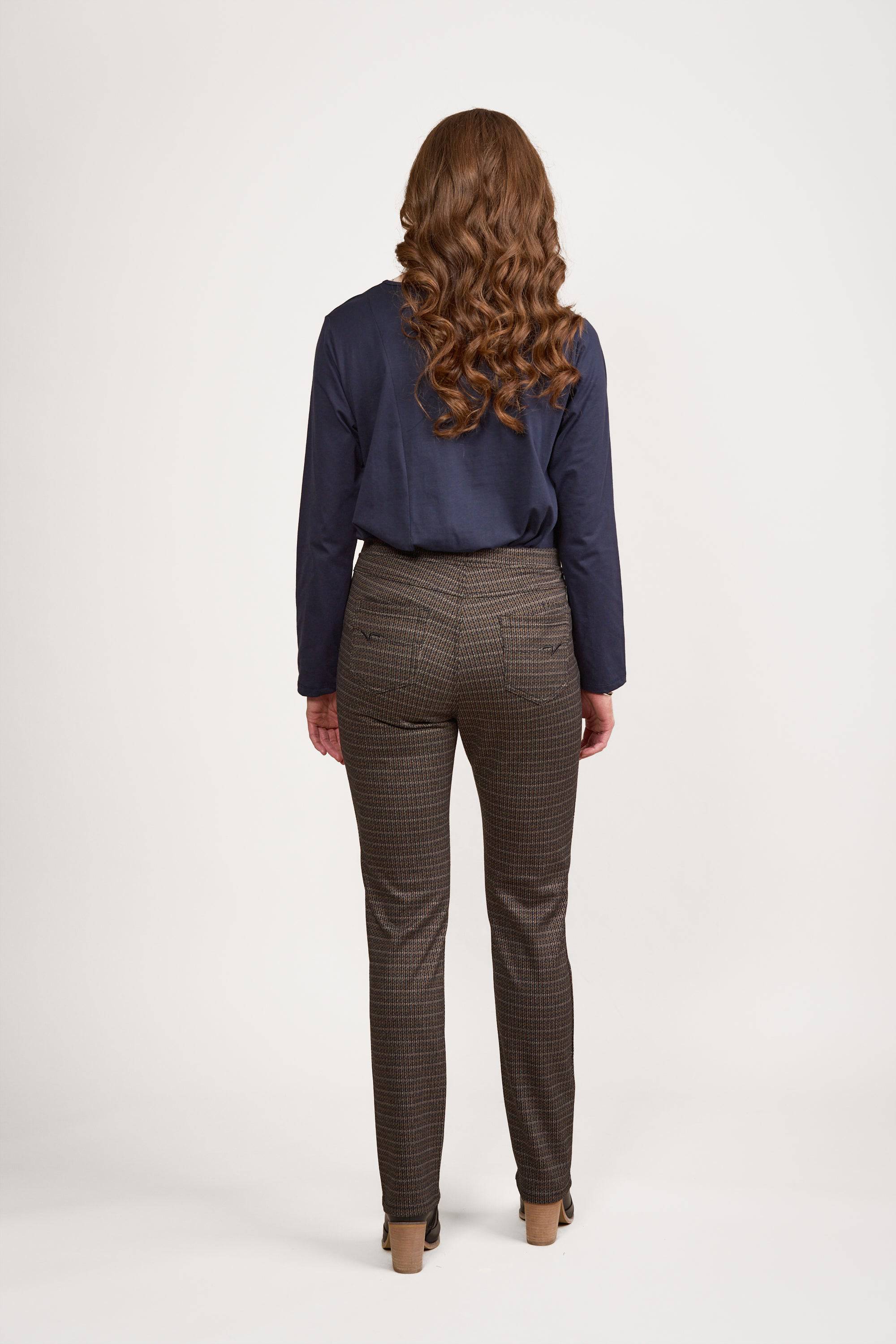 A woman with long, wavy brown hair faces away, wearing a navy Bay Knit Pull On - Sutton top by Vassalli, Vassalli Stretch Fabric Pants in a brown check pattern, and beige heels against a plain white background.