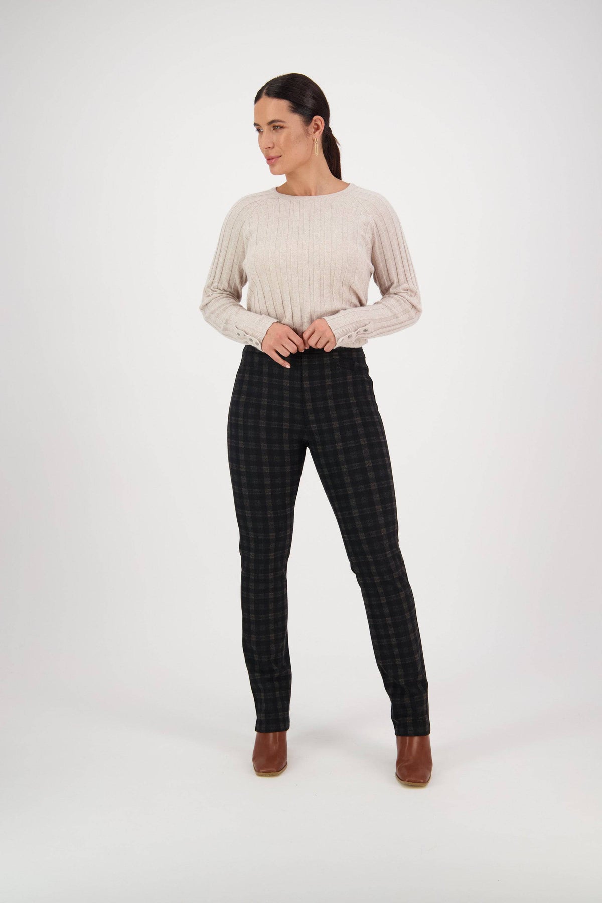A woman stands in a studio wearing a light beige ribbed sweater, Vassalli Bay Ponti Pull On - Halifax pants in dark plaid, and brown ankle boots. She looks to the side with her hands on her waistband against a plain white background.