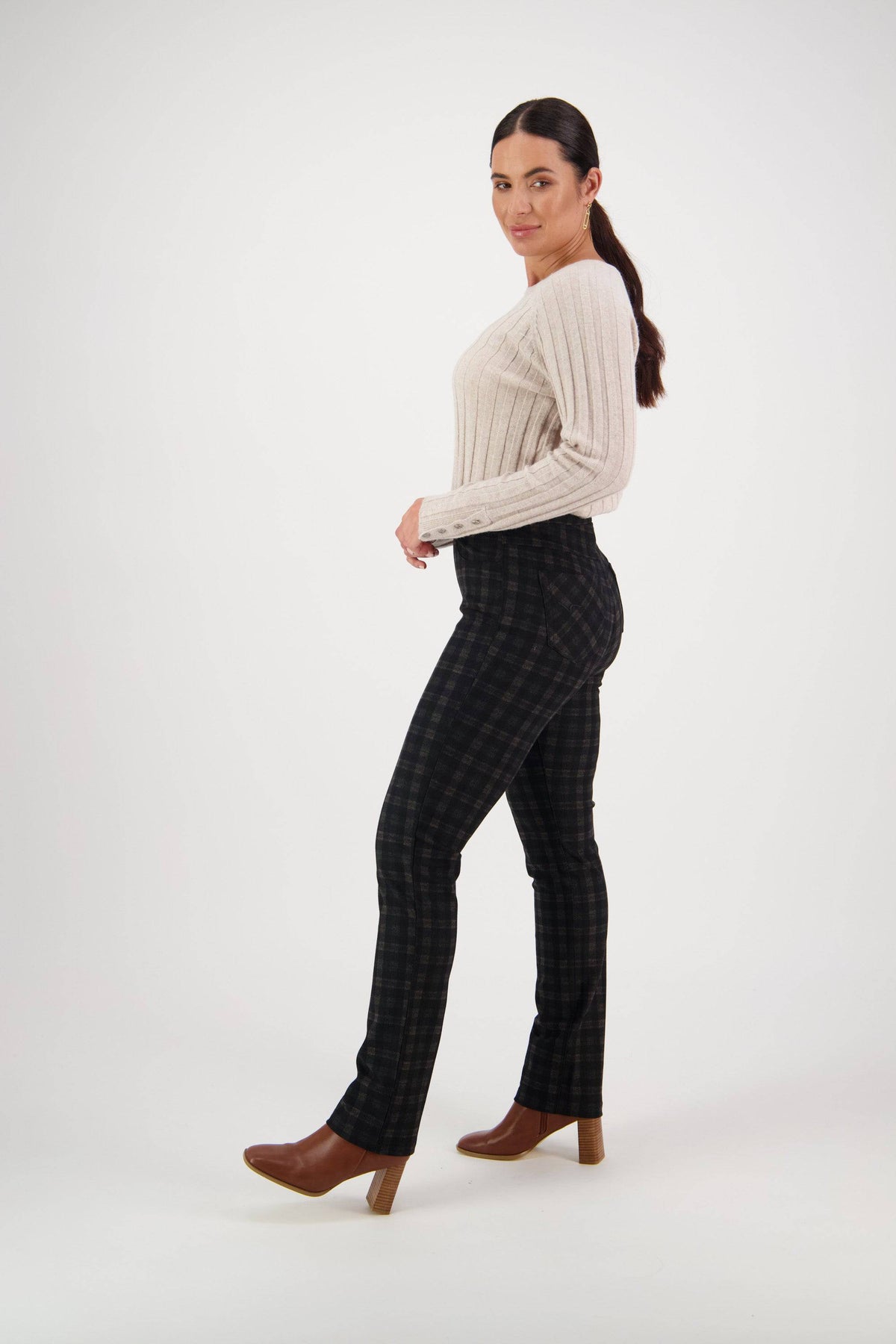 A woman with long dark hair in a ponytail poses sideways, wearing a cream ribbed sweater, Vassalli Bay Ponti Pull On - Halifax pants, and brown heeled boots, against a plain white background.