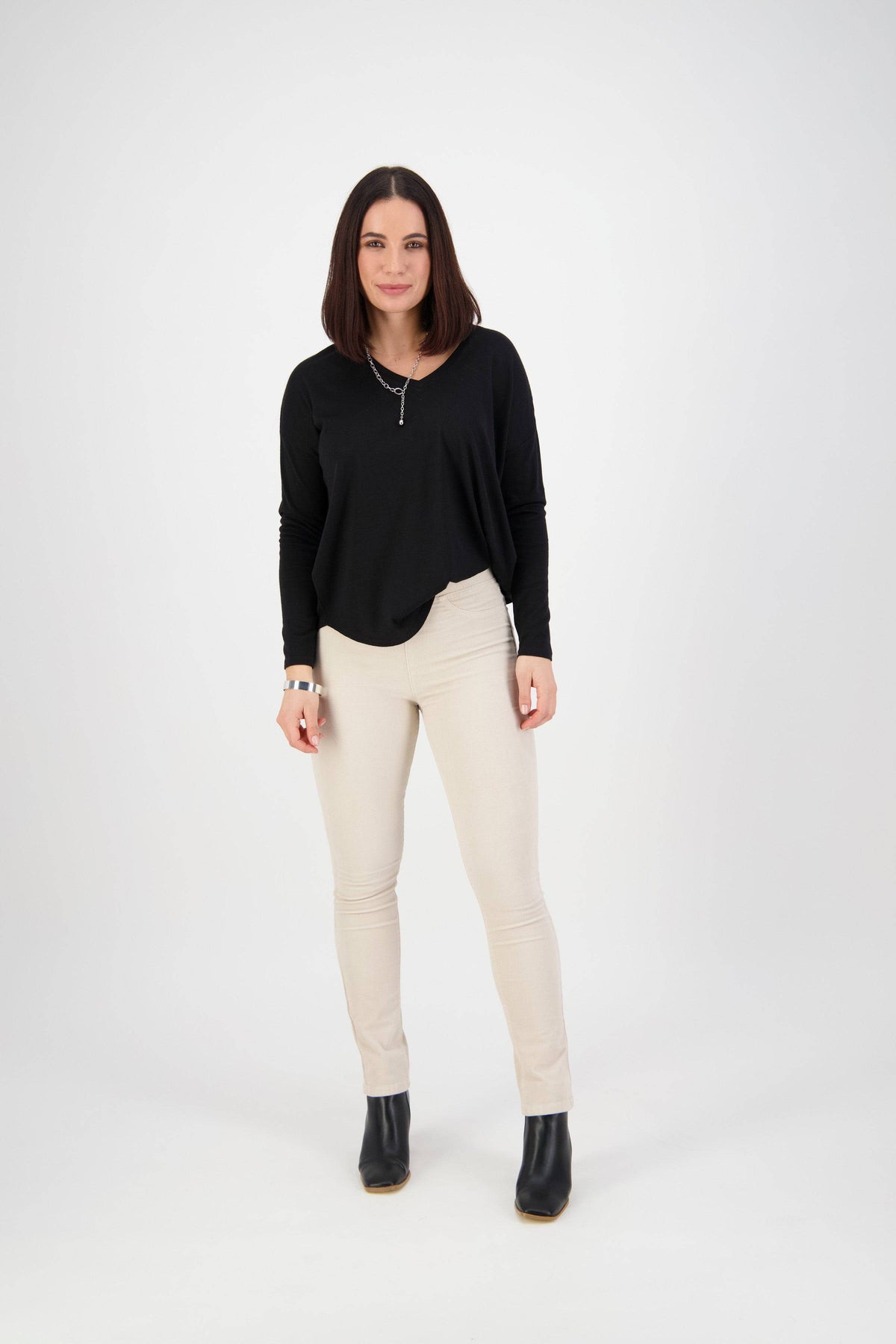 A woman with straight brown hair stands against a white background, wearing a black long-sleeve top, Bay Cord Pull On - Bone pants by Vassalli, black ankle boots, and a necklace, looking confidently at the camera.