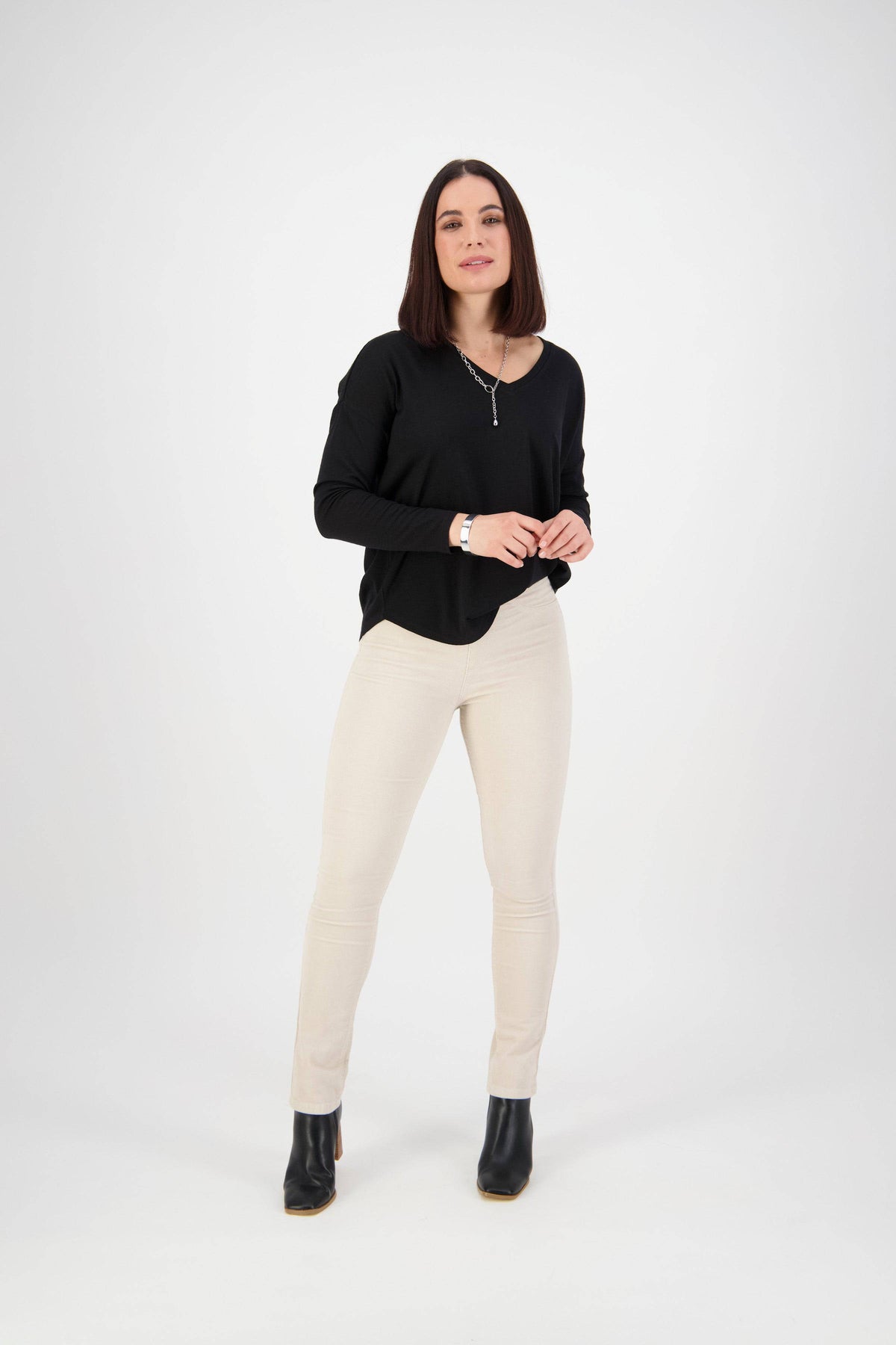 A woman with shoulder-length dark hair and a relaxed expression stands against a white background, wearing a black long-sleeve top and Vassalli Bay Cord Pull On pants in Bone, paired with black ankle boots.