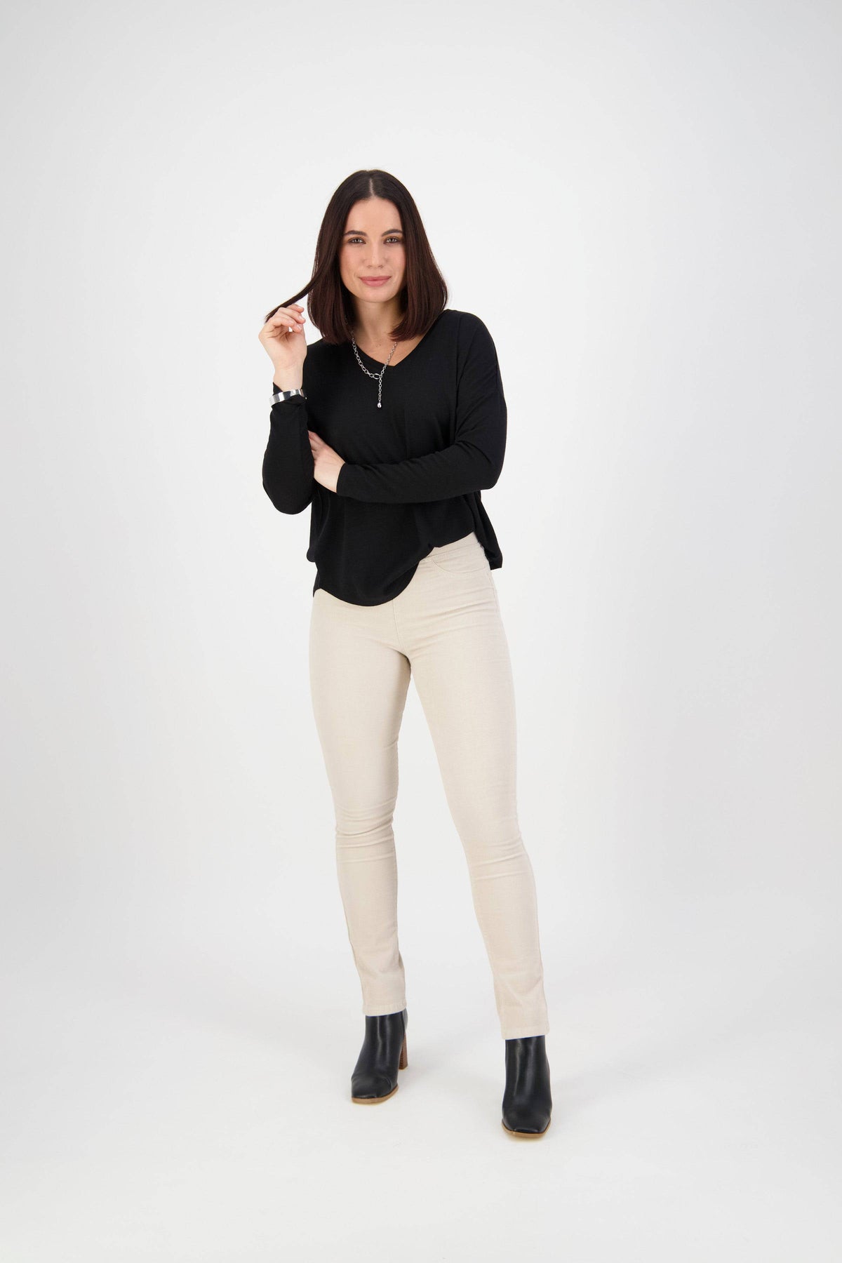 A woman with straight dark hair stands against a white background, wearing a black long-sleeve top, Vassalli Bay Cord Pull On pants in Bone, and black ankle boots. She smiles slightly, holding a strand of her hair with one hand.