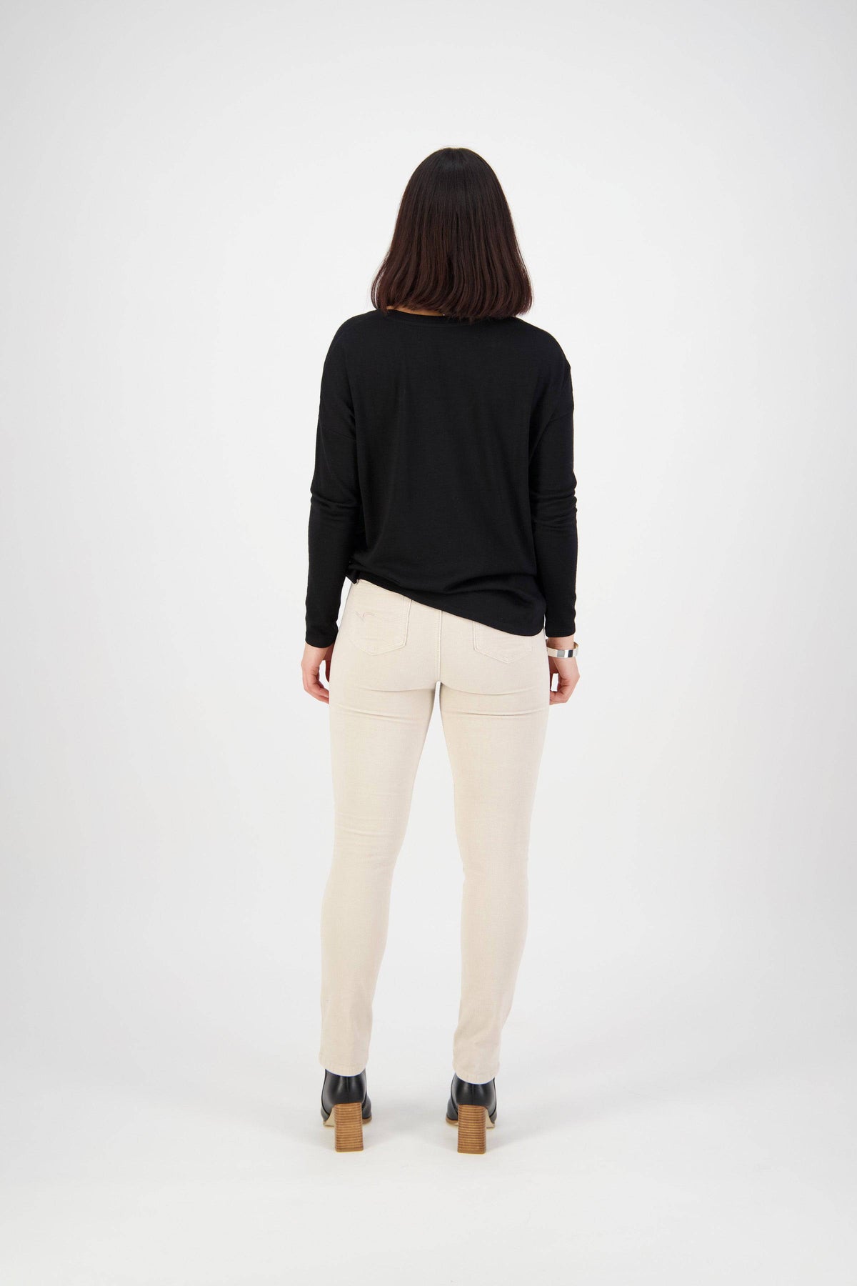 A person with shoulder-length dark hair stands facing away, wearing a black long-sleeve top, Vassalli Bay Cord Pull On pants in Bone, and black heeled shoes against a plain white background.