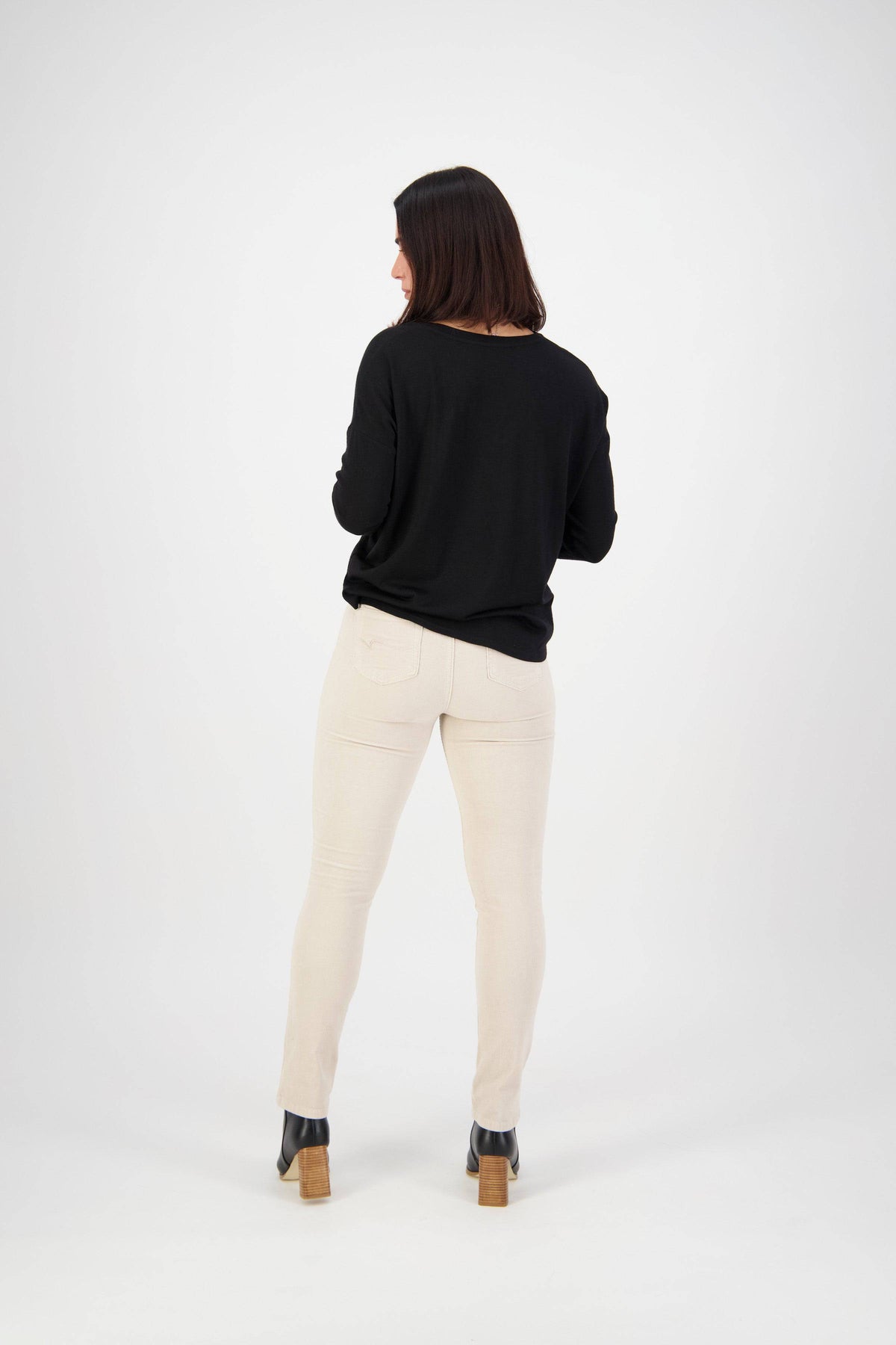 A woman with shoulder-length dark hair, facing away, wears a black long-sleeve top, Vassalli Bay Cord Pull On pants in Bone, and black heeled boots against a plain white background.