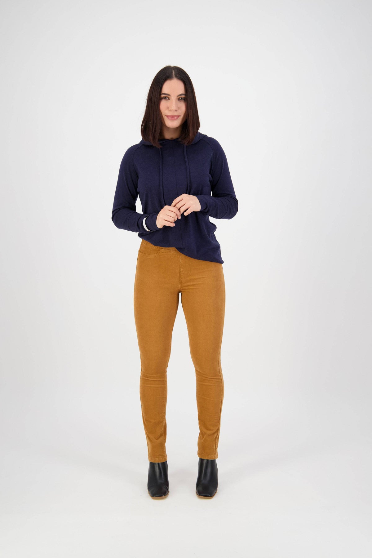 A woman with shoulder-length dark hair faces forward, wearing a navy hoodie, Vassalli Bay Cord Pull On pants in Copper, and black ankle boots against a plain white background.