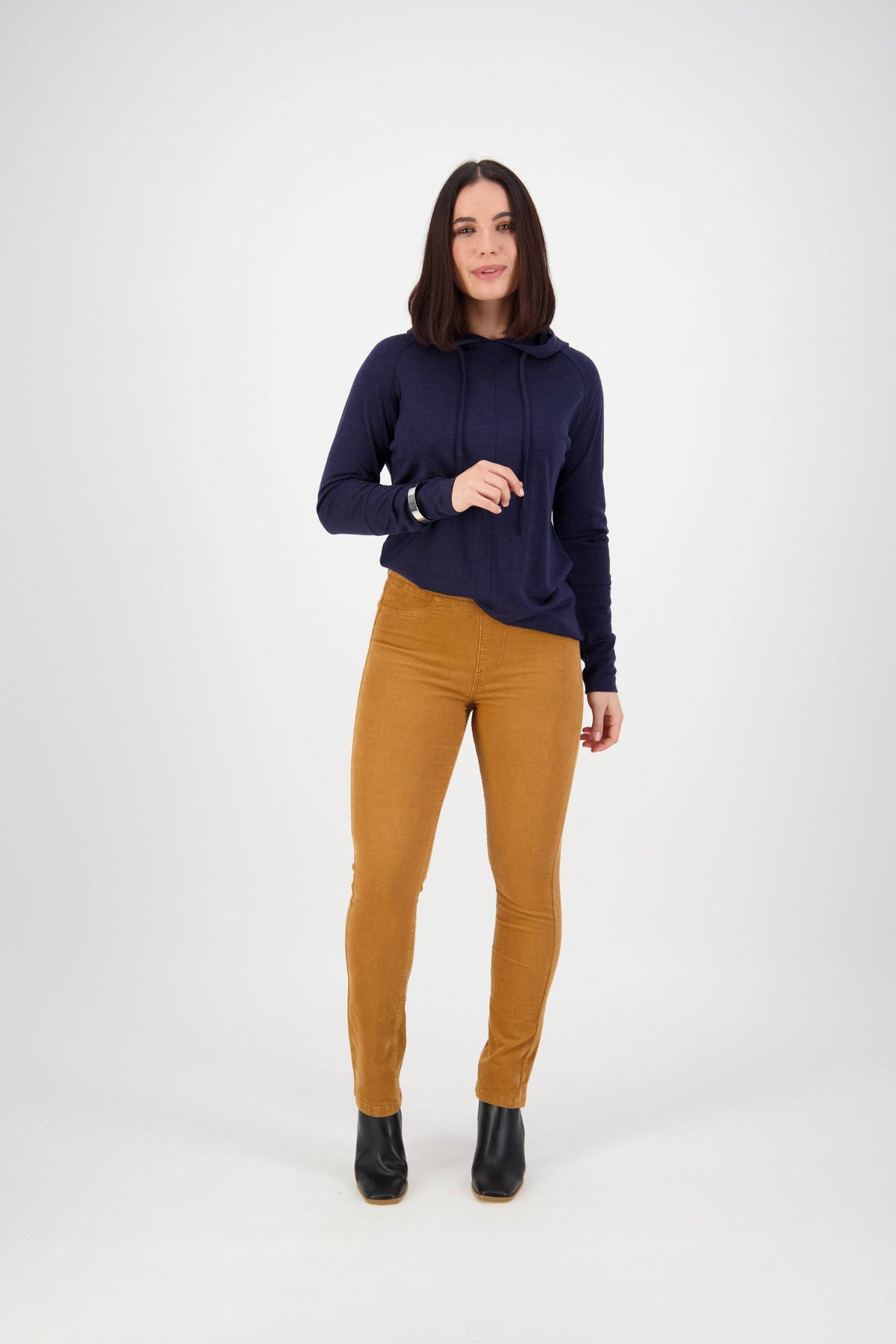 A woman with shoulder-length dark hair wears a navy hoodie, Vassalli Bay Cord Pull On pants in copper, and black ankle boots, standing against a plain white background.