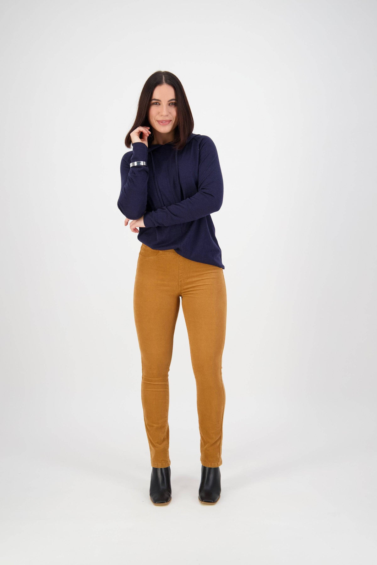 A woman with straight dark hair stands against a plain white background, smiling in a navy long-sleeve top, Vassalli Bay Cord Pull On pants in copper, and black ankle boots. She touches her chin with one hand.