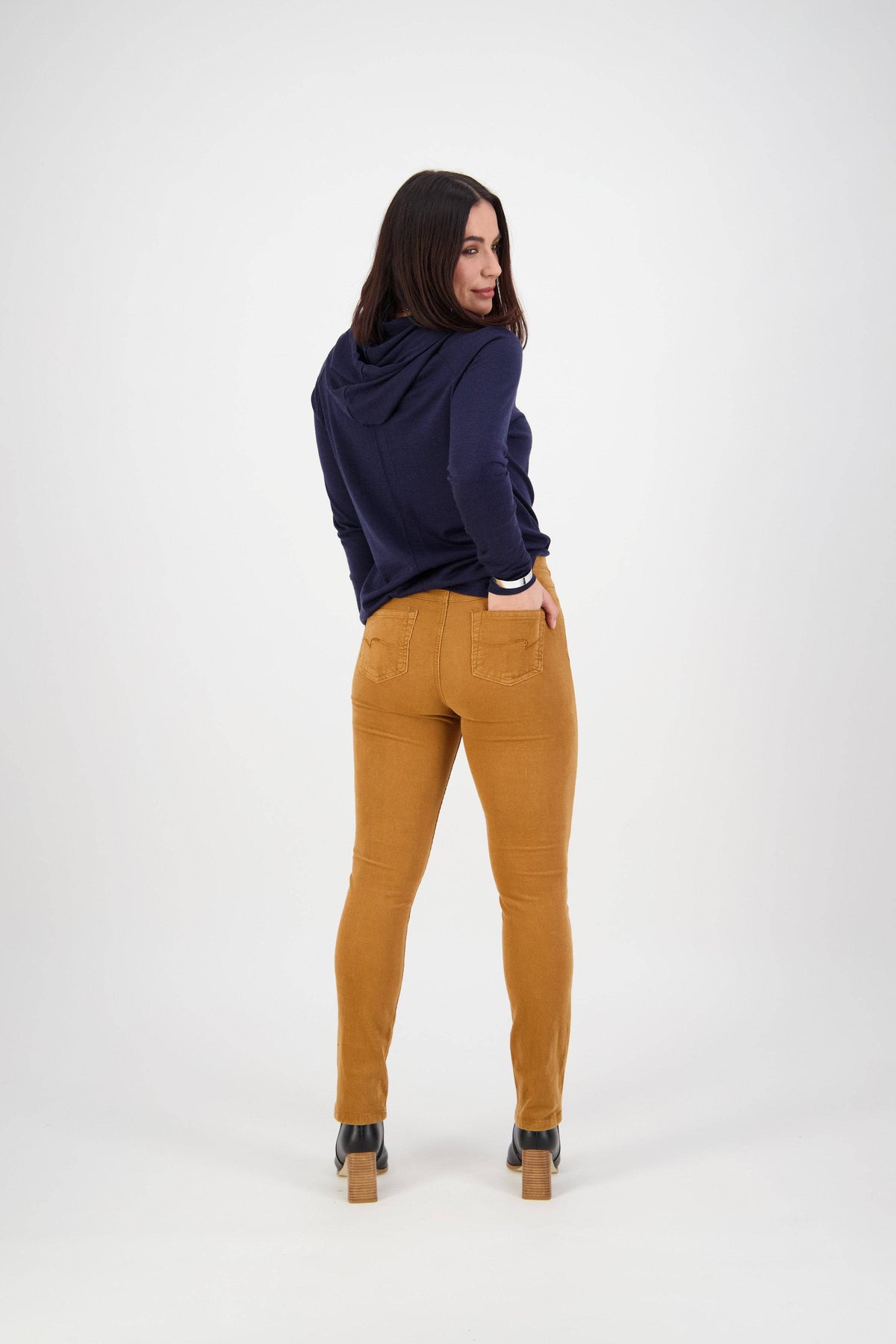 A woman faces away, glancing over her shoulder in a navy hoodie, Vassalli Bay Cord Pull On pants in copper, and heeled shoes. The background is plain white.