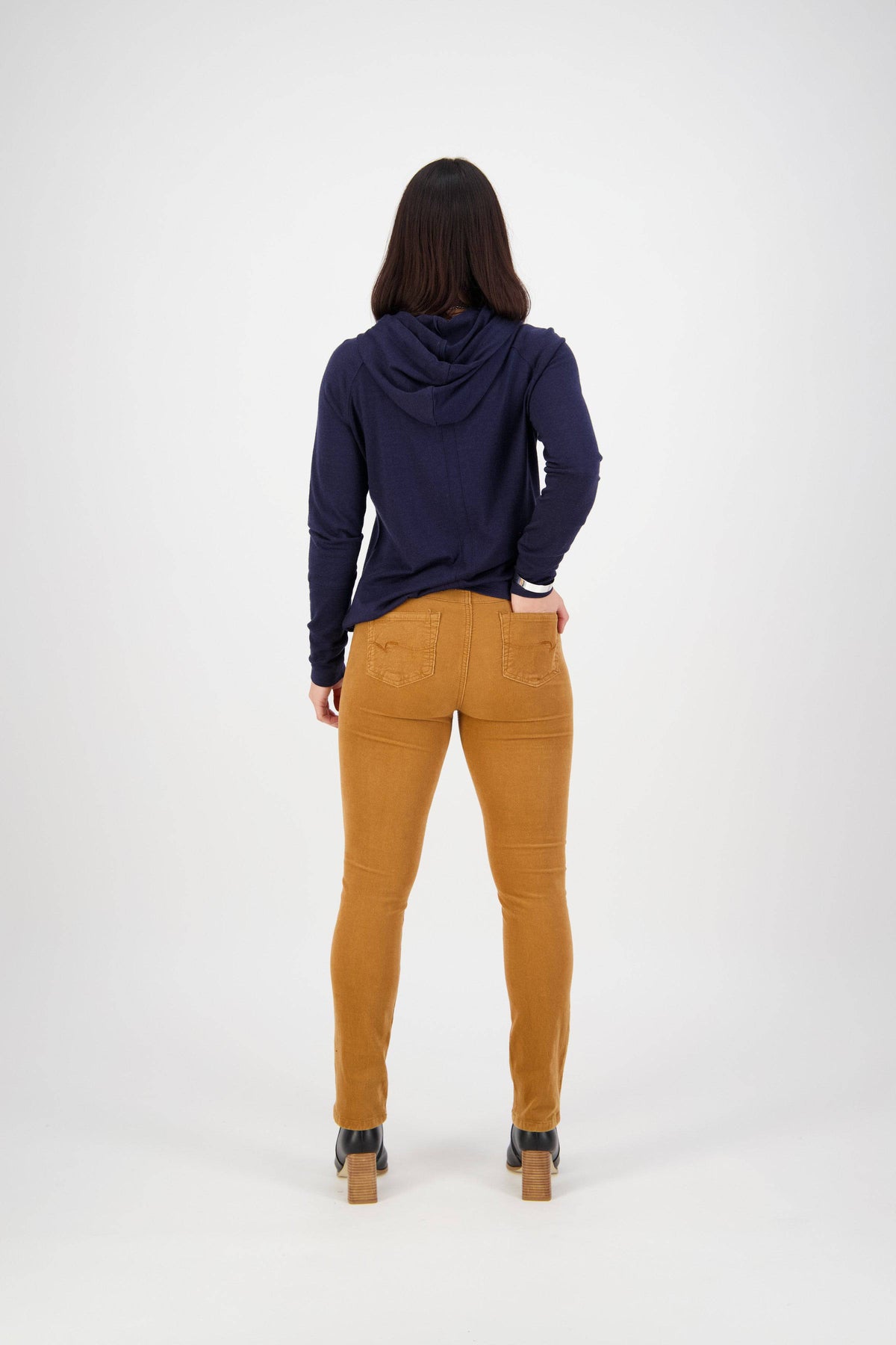 A person stands facing away from the camera, wearing a navy hooded long-sleeve shirt and Vassalli Bay Cord Pull On pants in Copper with heeled shoes, set against a plain white background.