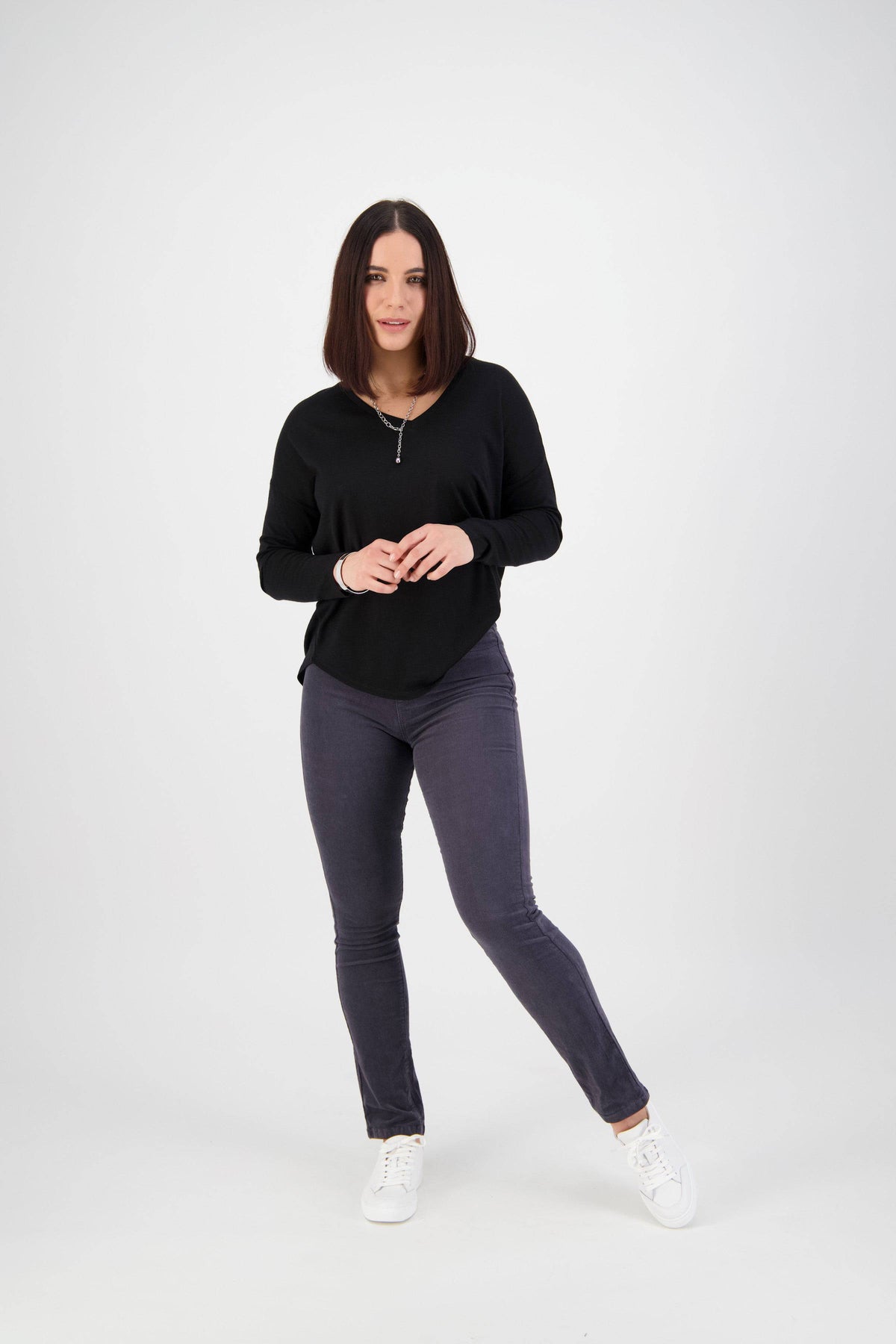 A woman with shoulder-length dark hair stands against a white background, wearing the Vassalli Bay Cord Pull On - Graphite slim leg pants, a black long-sleeve top, and white sneakers. She faces forward with a slight smile and relaxed posture.