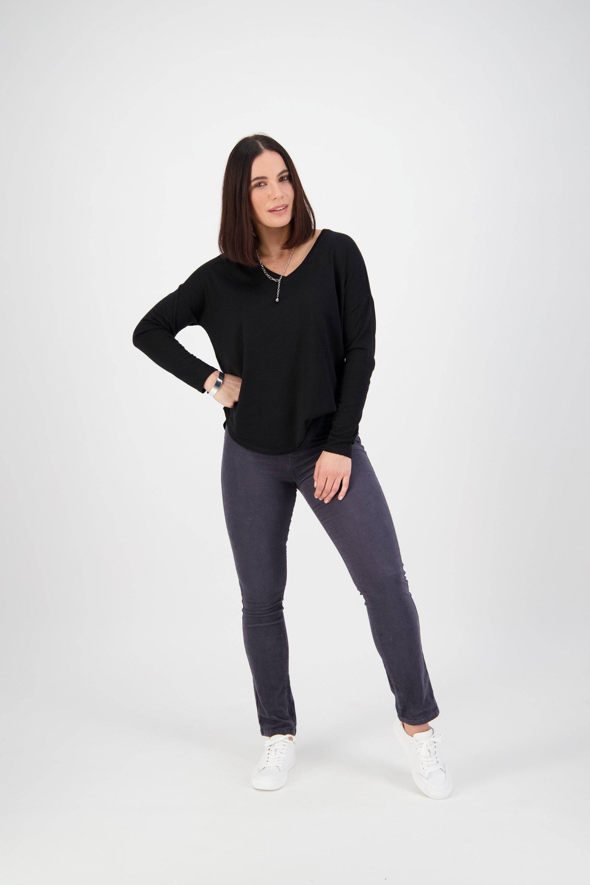 A woman with straight dark hair stands against a white background, wearing Vassalli's Bay Cord Pull On in Graphite and a black long-sleeve top. She smiles softly, one hand on her hip, the other relaxed by her side.