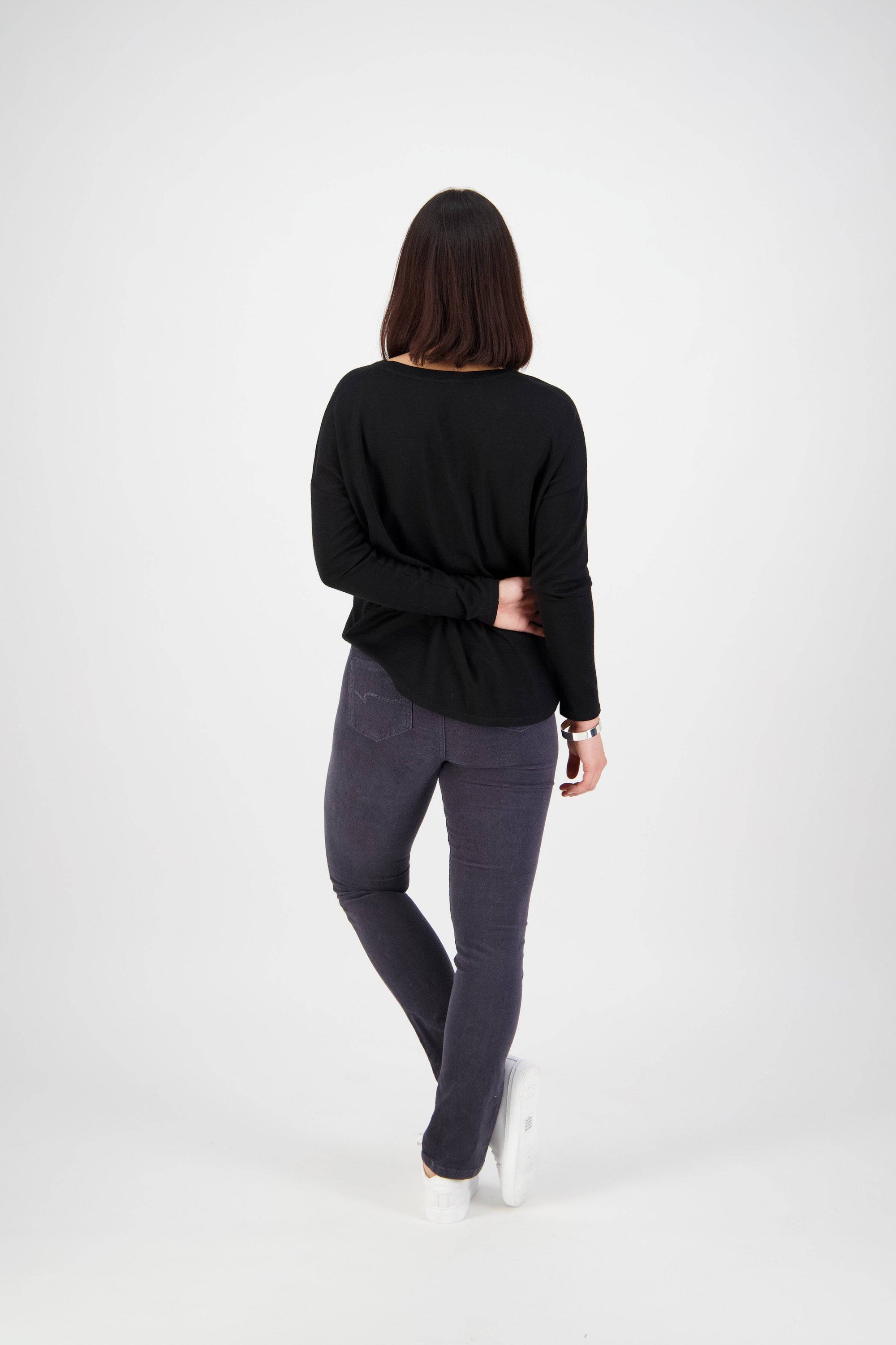 A person with shoulder-length dark hair faces away against a white background, wearing a black long-sleeve top, white shoes, and Vassalli Bay Cord Pull On - Graphite slim leg pants with an elastic waistband, one hand on the opposite arm.