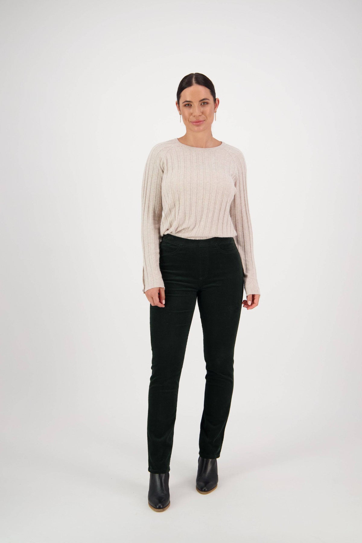 A woman stands facing forward against a plain white background, wearing a light beige long-sleeve sweater, Vassalli Bay Cord Pull On - Olive pants with a slim leg cut, and black ankle boots. Her dark hair is pulled back and she smiles gently.