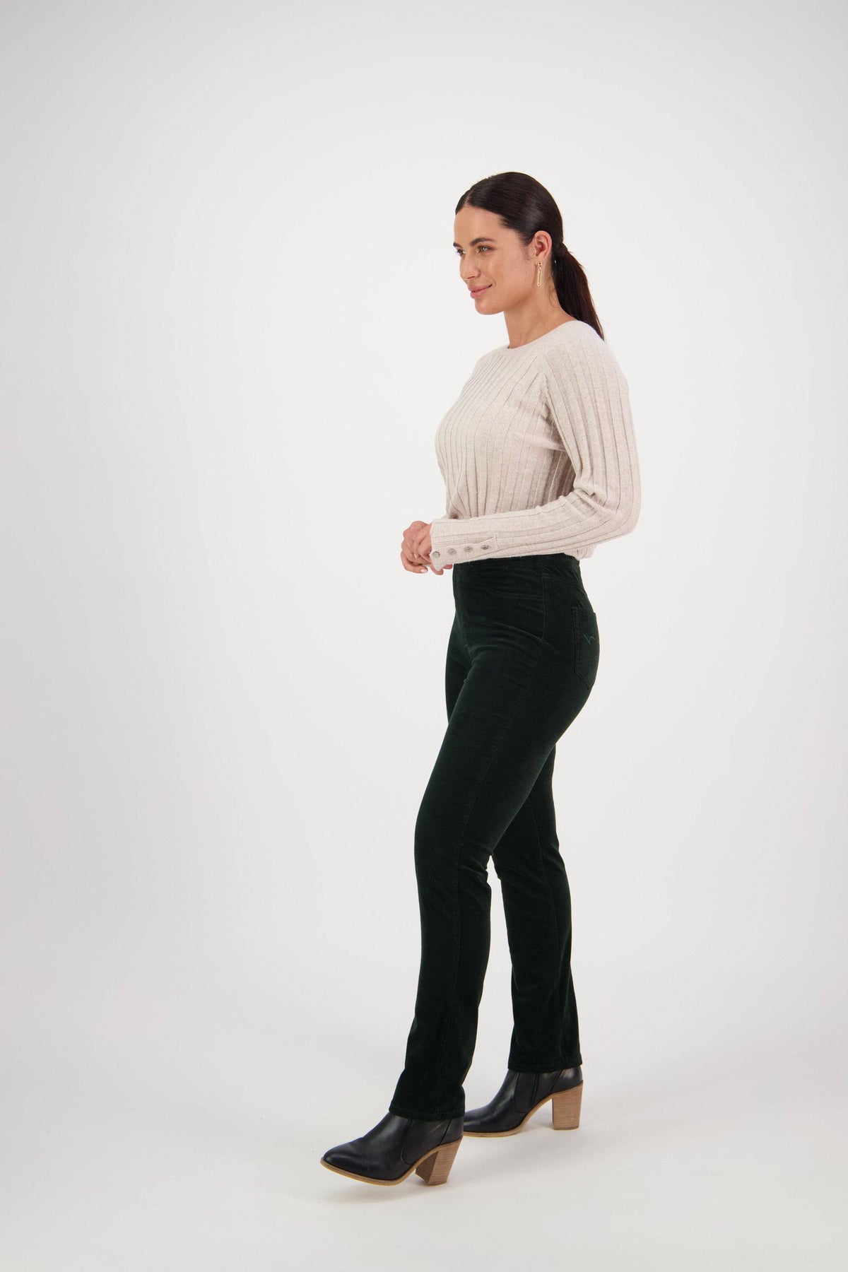 A woman stands in profile against a plain white background, wearing a cream ribbed sweater, Vassalli Bay Cord Pull On pants in Olive, and heeled boots. Her dark hair is pulled back in a low ponytail.