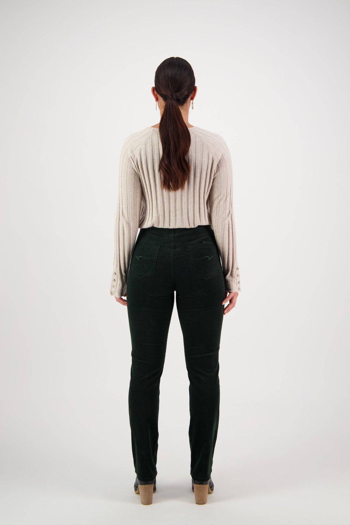 A woman stands facing away, wearing a beige ribbed sweater, Vassalli Bay Cord Pull On pants in olive, and heeled shoes. Her hair is styled in a low ponytail against a plain white background.