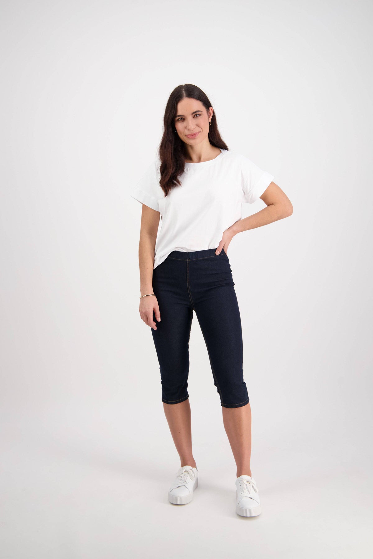A woman with long brown hair stands against a white background, wearing the Vassalli Atlas 3/4 Pant in Indigo Denim (Contrast Stitch), a white t-shirt, and white sneakers. One hand is on her hip and she looks relaxed.