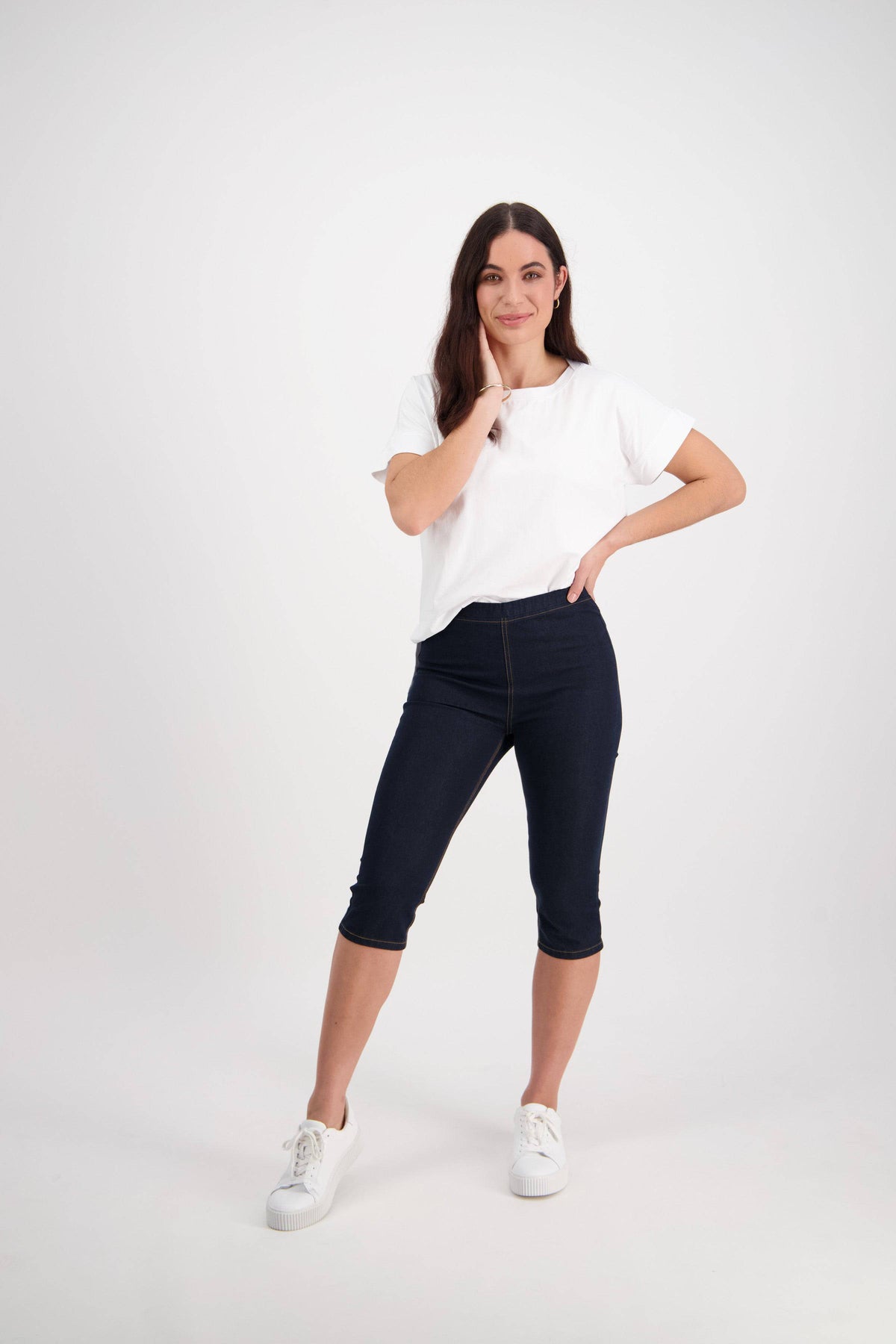 A woman with long brown hair wearing a white tee, white sneakers, and Vassalli Atlas 3/4 Pant - Indigo Denim (Contrast Stitch) stands confidently against a plain white background, one hand on her hip and the other touching her face.