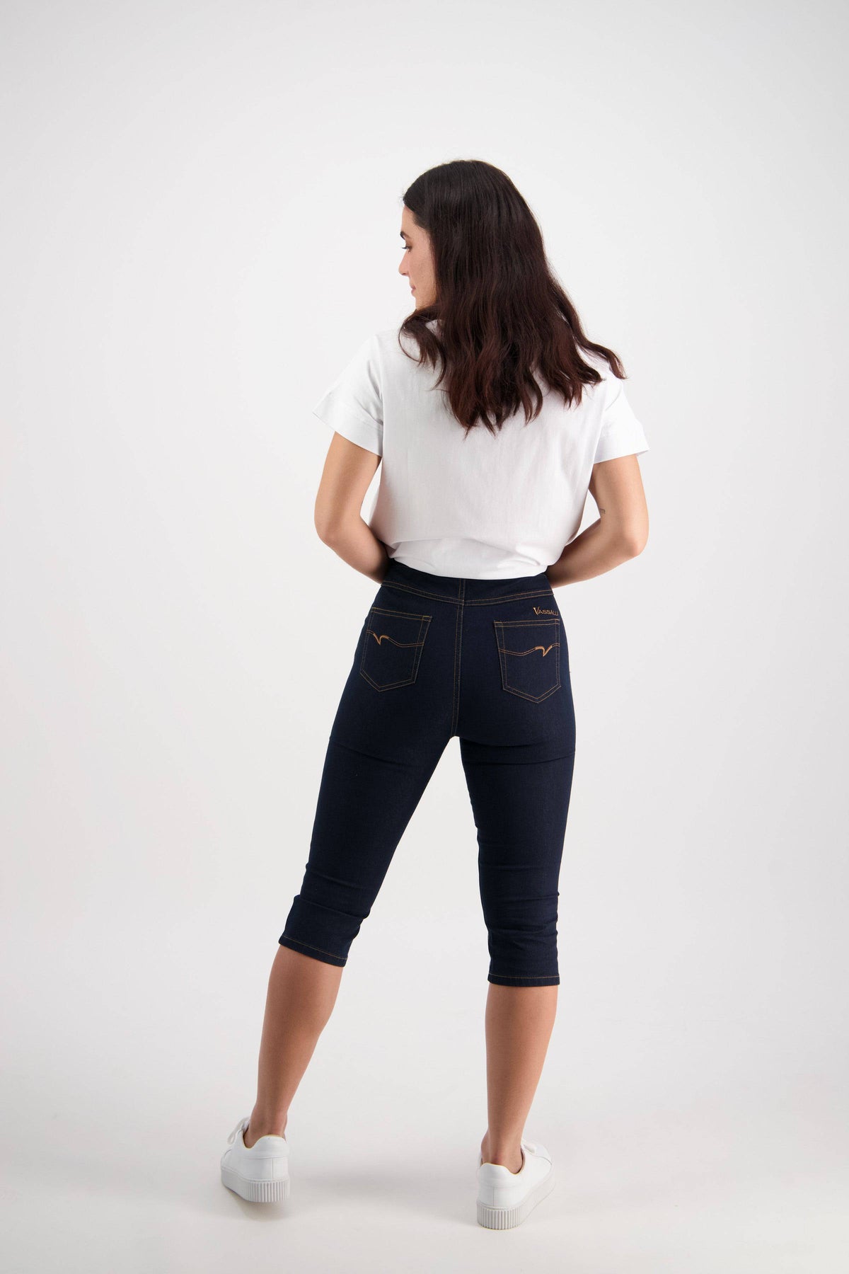 A woman with long dark hair, wearing a white t-shirt and Vassalli Atlas 3/4 Pant - Indigo Denim (Contrast Stitch), stands facing away from the camera against a plain white background.