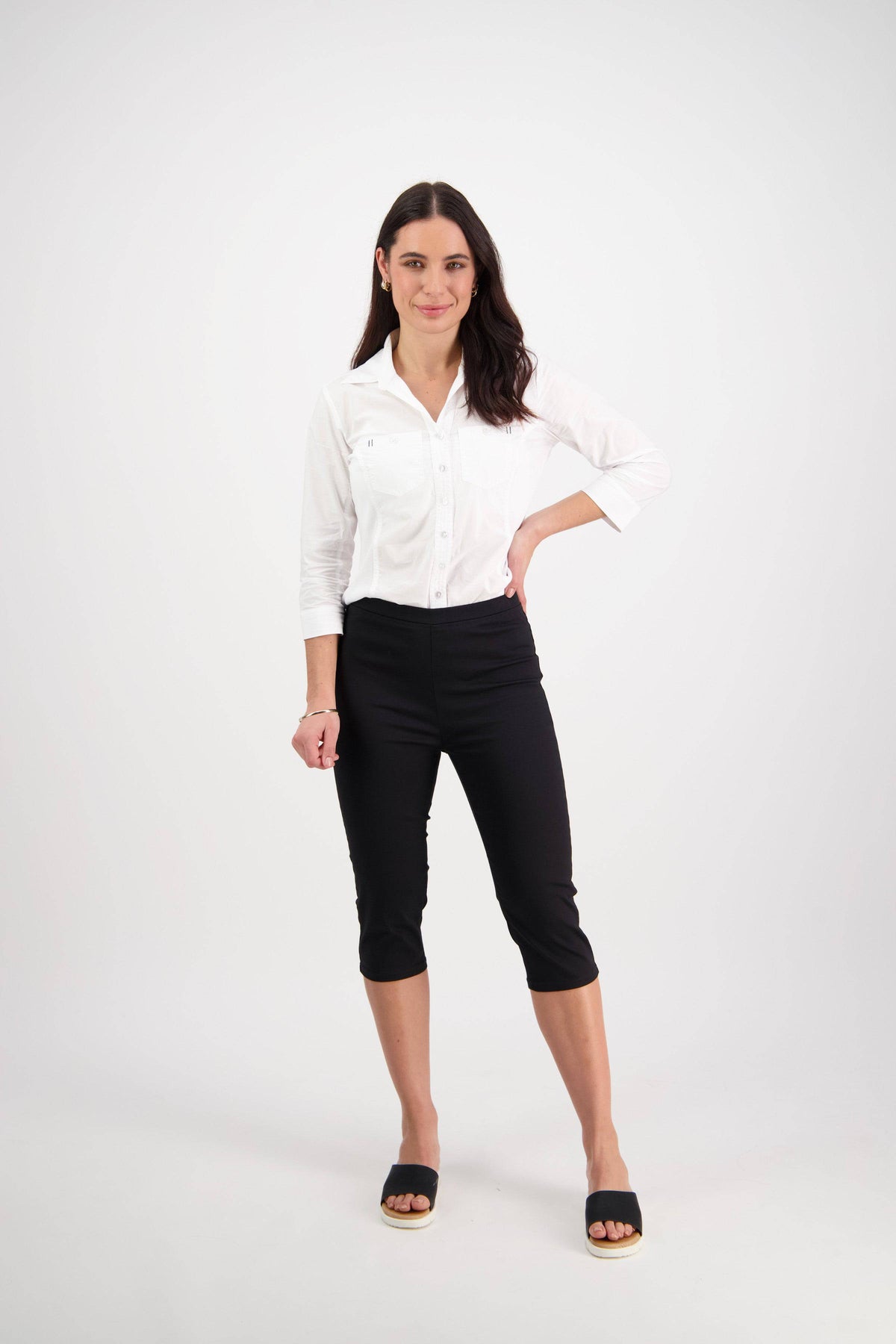 A woman with long dark hair stands confidently against a white background, wearing Vassalli’s Atlas 3/4 Pant in black, paired with a white button-up shirt and black slide sandals, exuding a relaxed, friendly vibe.