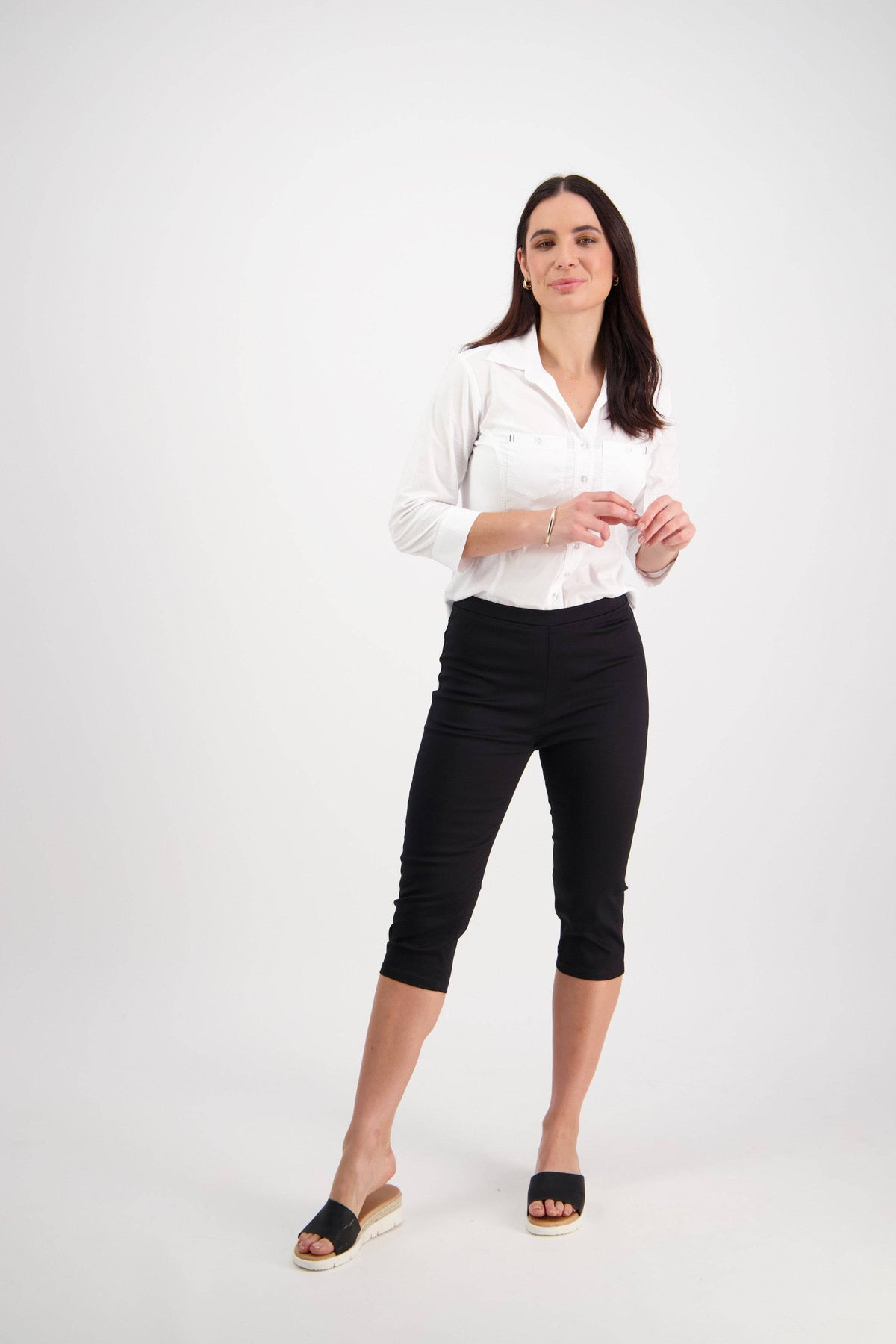 A woman with long dark hair stands against a plain white background, gently smiling as she wears the Vassalli Atlas 3/4 Pant in black and a white button-up shirt, paired with black slide sandals and her hands loosely held in front of her.