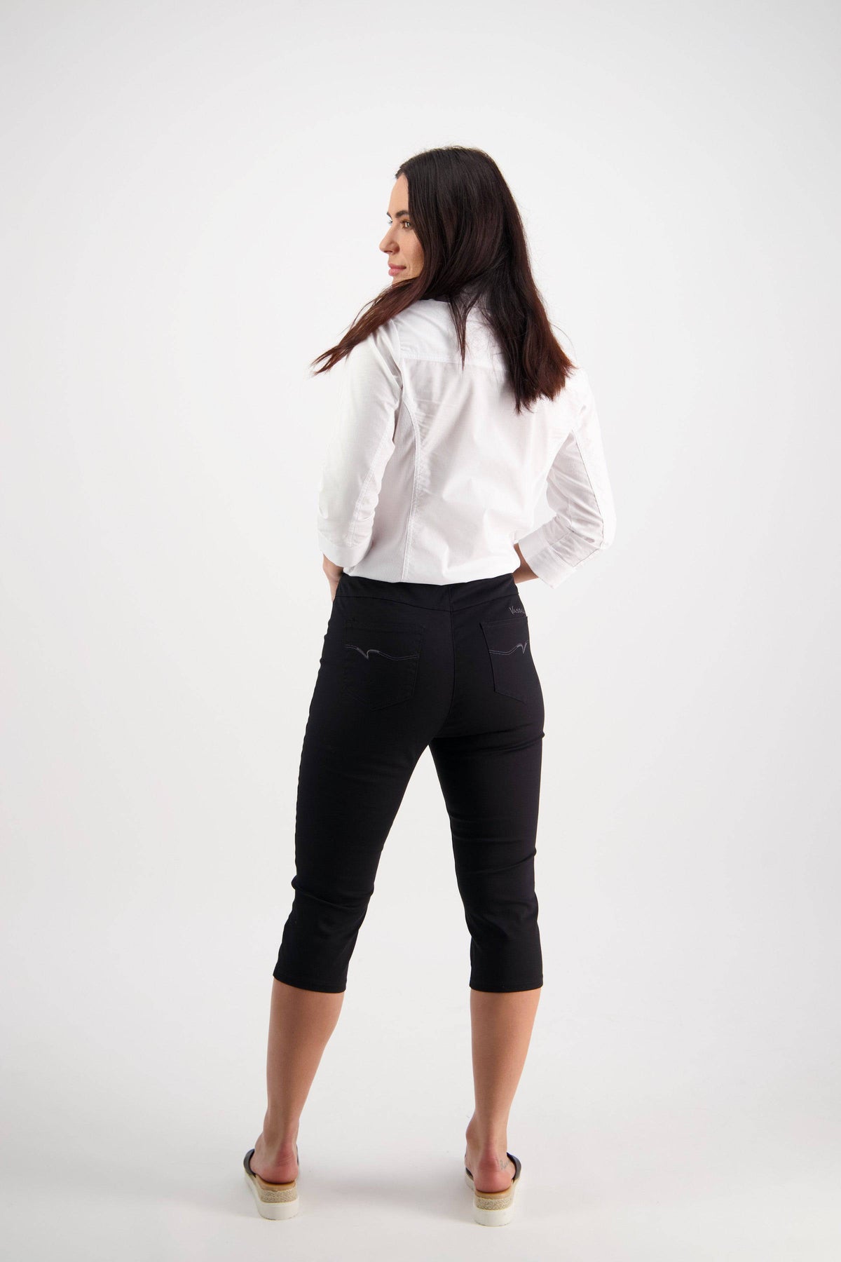 A woman with long dark hair faces away, looking over her left shoulder. She wears a white blouse and Vassalli Atlas 3/4 Pant in black with an elastic waistband, paired with beige sandals against a plain white background.