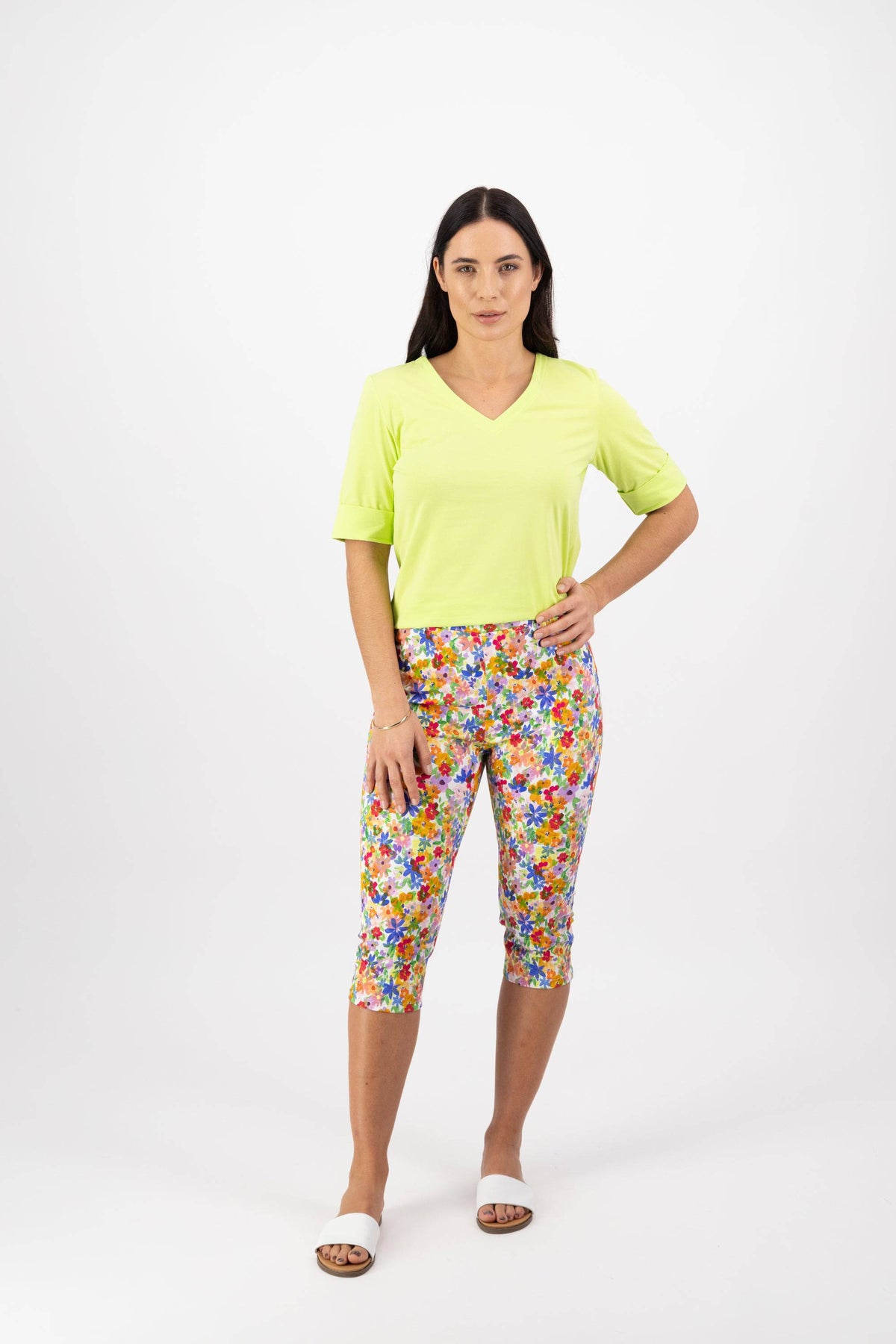 A woman with long dark hair and a neutral expression stands against a white background in a light green V-neck top and Vassalli Atlas 3/4 Pant - Bloom, paired with white slide sandals. One hand rests on her hip.