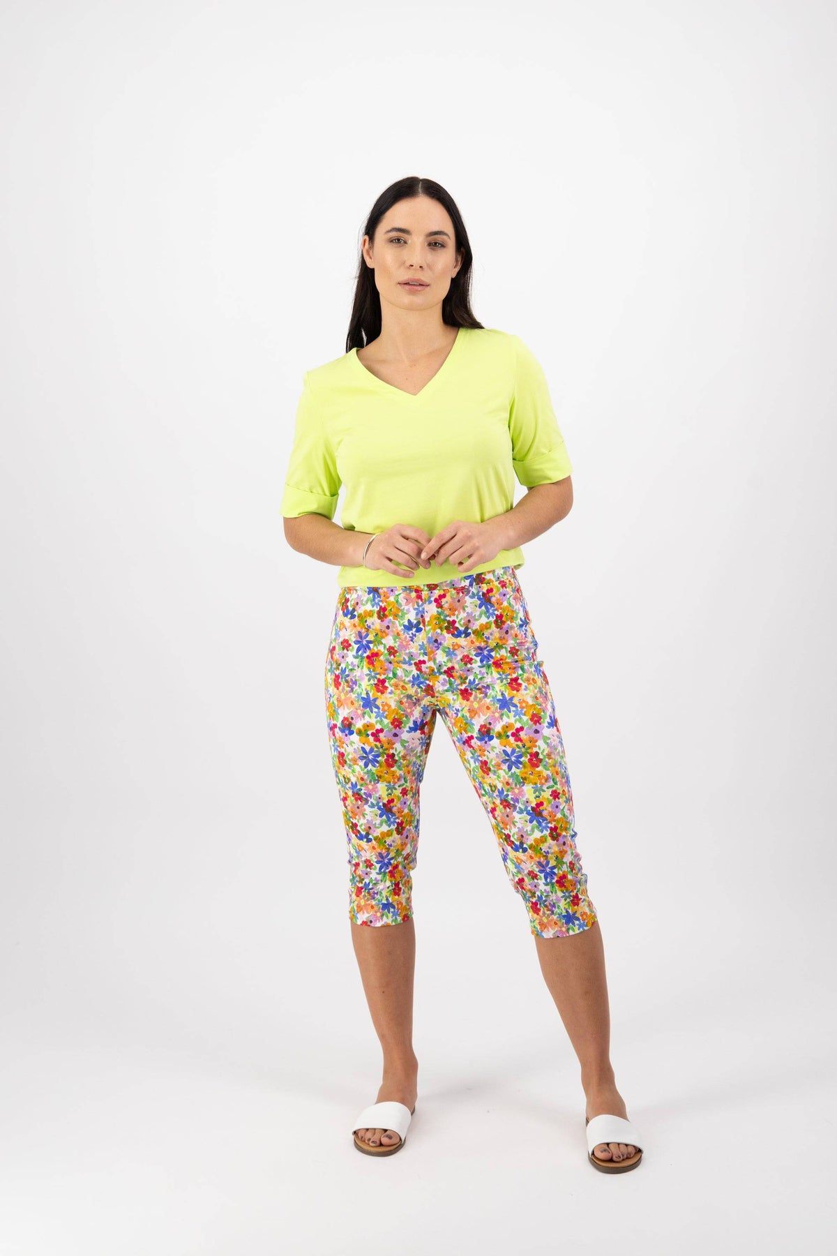 A woman with long dark hair and a neutral expression stands against a white background wearing a light green V-neck top, Vassalli Atlas 3/4 Pant in Bloom print, and white slide sandals.