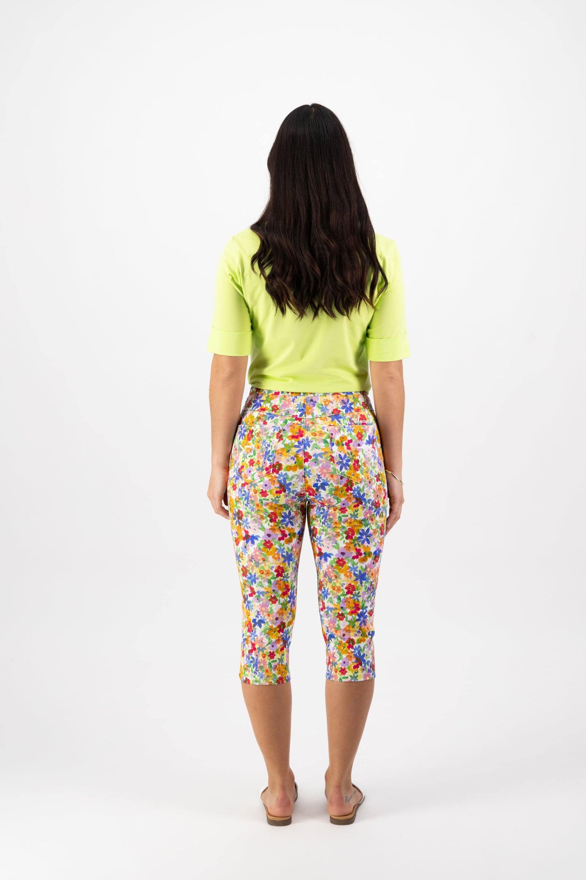 A woman with long dark hair faces away, wearing a light green short-sleeve top and Vassalli Atlas 3/4 Pant - Bloom with sandals, set against a plain white background.