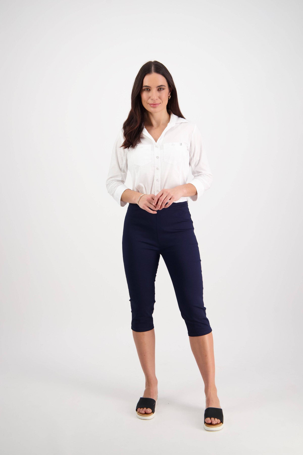 A woman with long dark hair and a neutral expression stands against a white background, wearing the Vassalli Atlas 3/4 Pant in navy, a white long-sleeve button-up shirt, and black slide sandals.