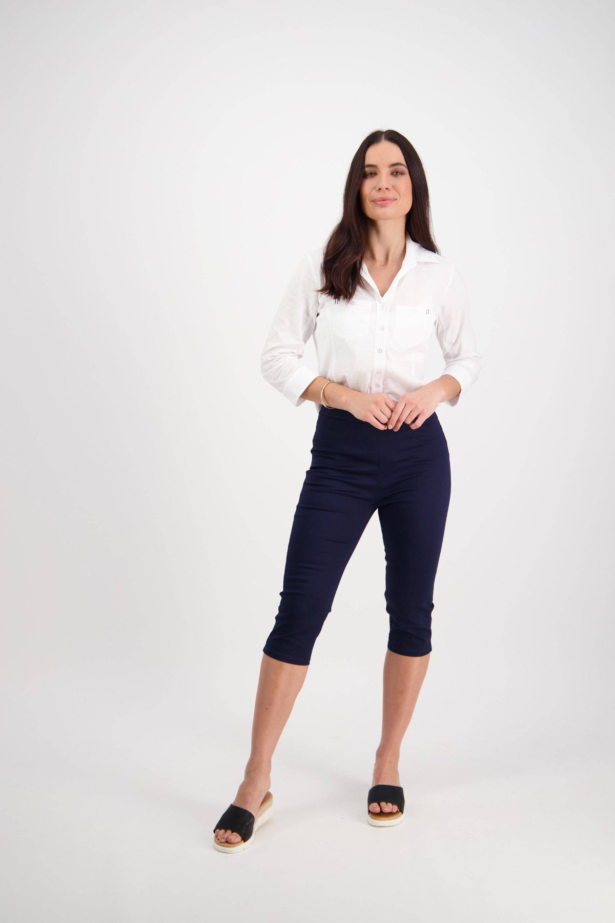 A woman poses confidently against a white background, wearing the Vassalli Atlas 3/4 Pant in navy with an elastic waistband, a white button-up shirt, and black open-toe sandals. Her long brown hair adds to the relaxed look.