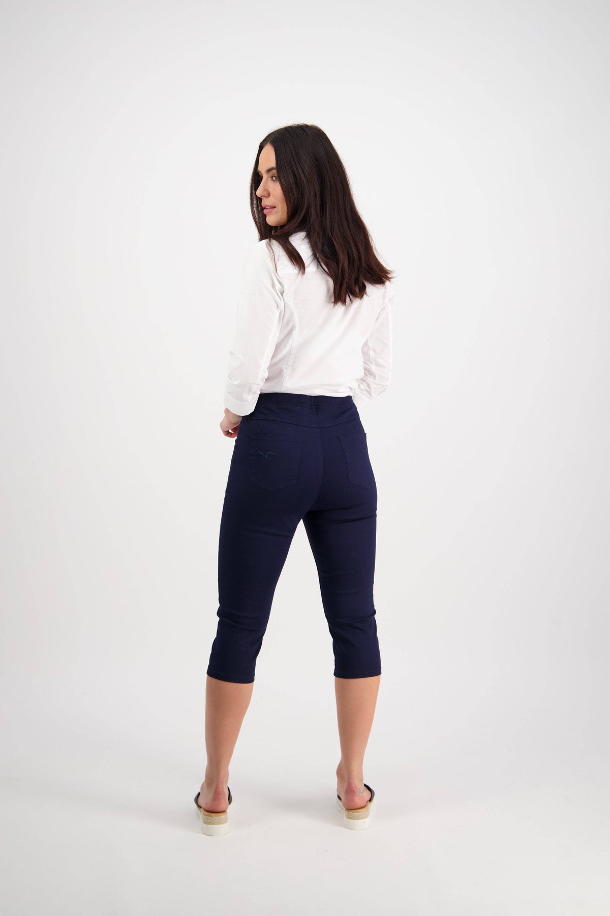 A woman with long dark hair faces away, wearing a white long-sleeve shirt, Vassalli's Atlas 3/4 Pant in navy with an elastic waistband, and beige sandals against a plain white background.