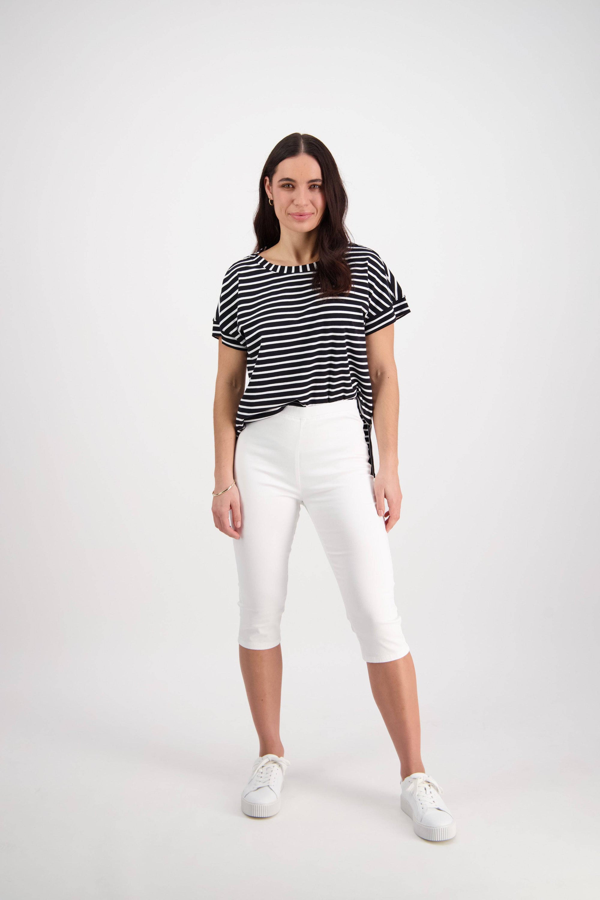 A woman with long brown hair stands confidently against a white background, wearing Vassalli's Atlas 3/4 Pant in white, a black and white striped top, and white sneakers.