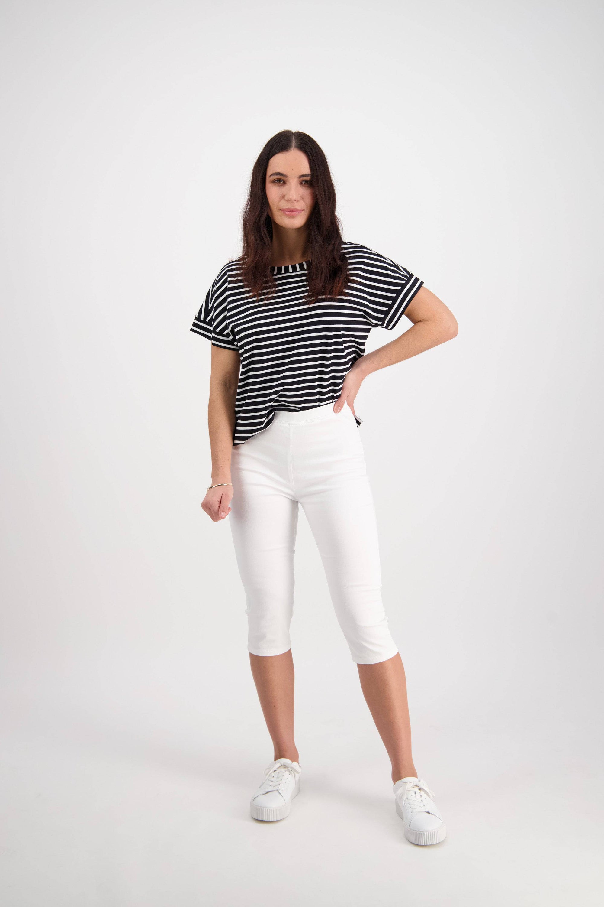 A woman with long brown hair poses against a white background, wearing the Vassalli Atlas 3/4 Pant in White, a black and white striped tee, and white sneakers. She stands with one hand on her hip, looking at the camera.