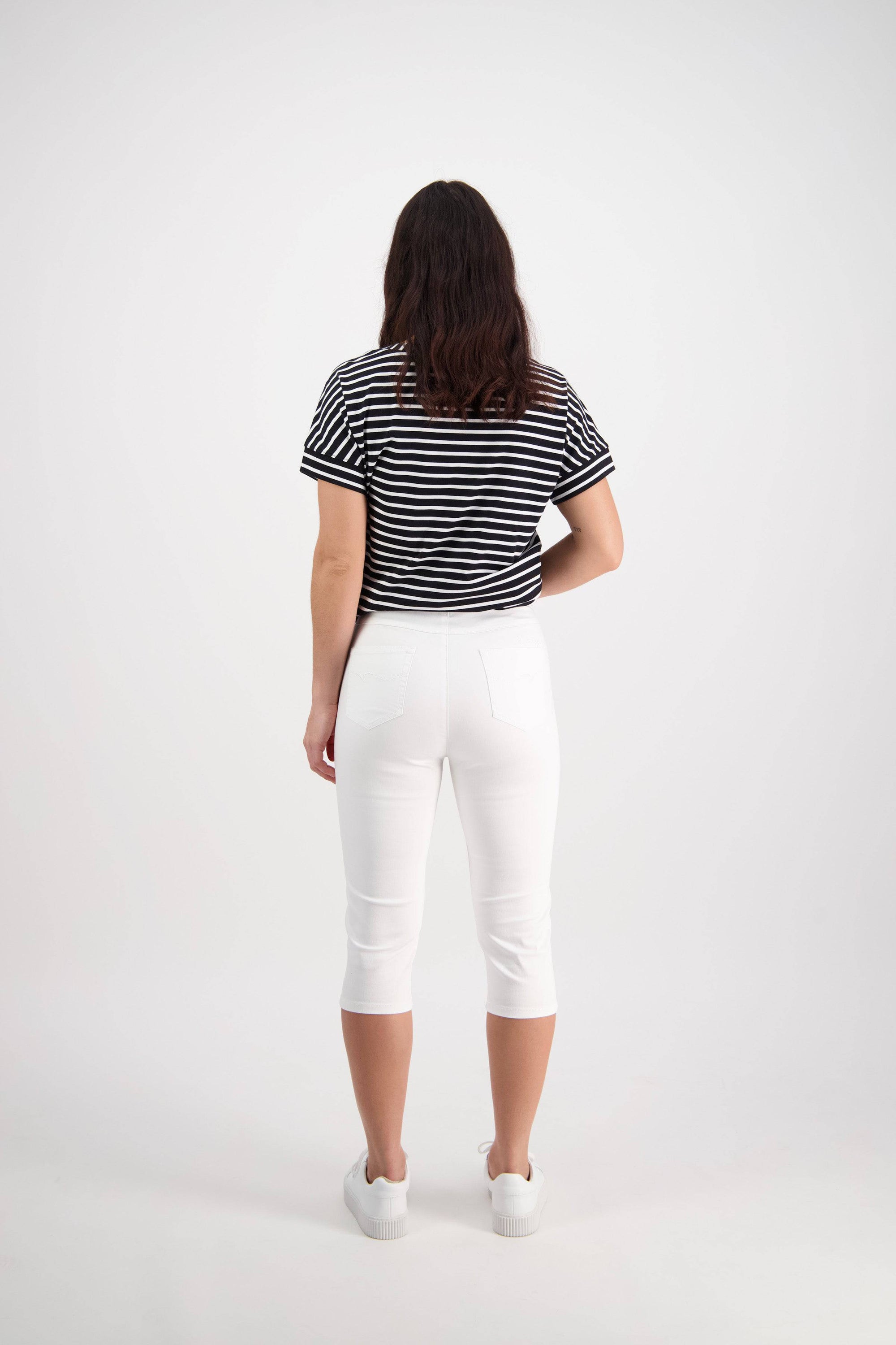 A woman stands facing away, wearing a black and white striped short-sleeve shirt, Vassalli Atlas 3/4 Pant in white with a skinny leg fit, and white shoes, against a plain white background.