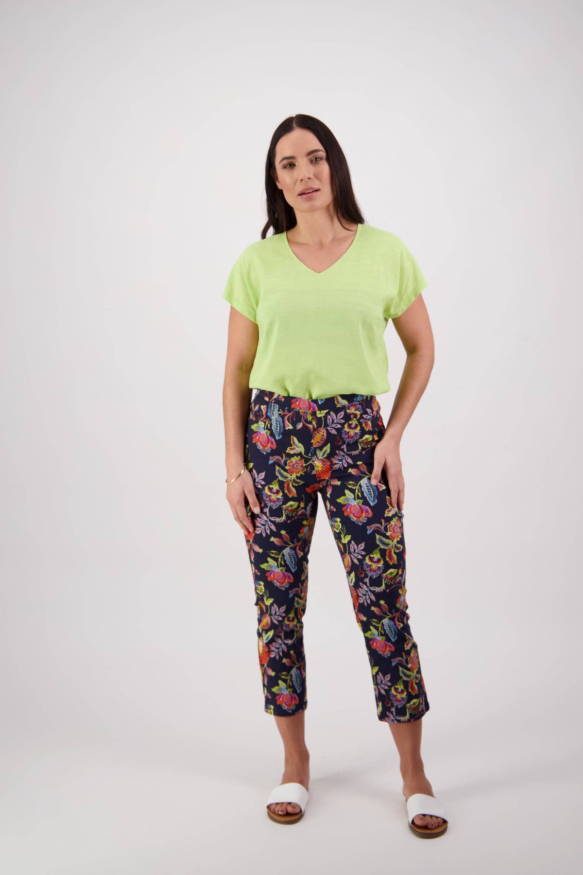 A woman with long dark hair and a relaxed posture stands against a white background, wearing Vassalli's Sierra 7/8 Pant - Brazil, a light green short-sleeve top, and white open-toe sandals, with one hand in her pocket.