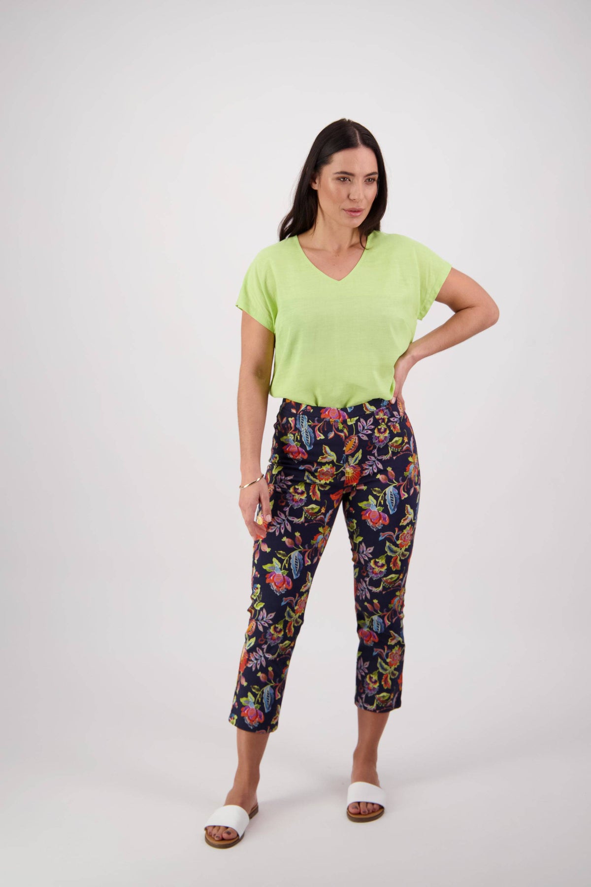 A woman with long dark hair stands against a white background, wearing the Vassalli Sierra 7/8 Pant - Brazil, a light green short-sleeve top, and white slide sandals. She poses with one hand on her hip and a neutral expression.